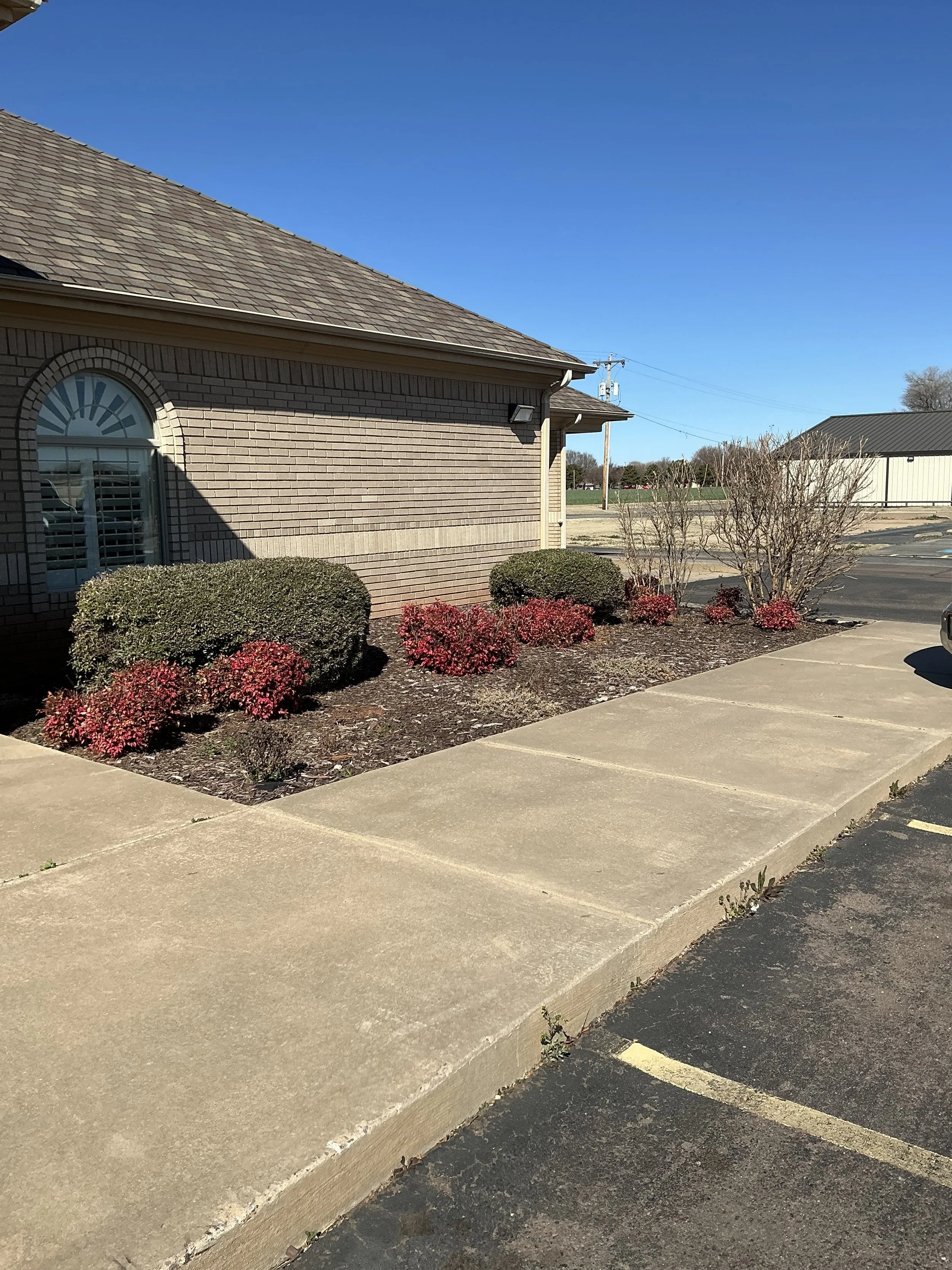 A sidewalk in front of a brick building with bushes and small trees, a parking lot, and a clear blue sky.
