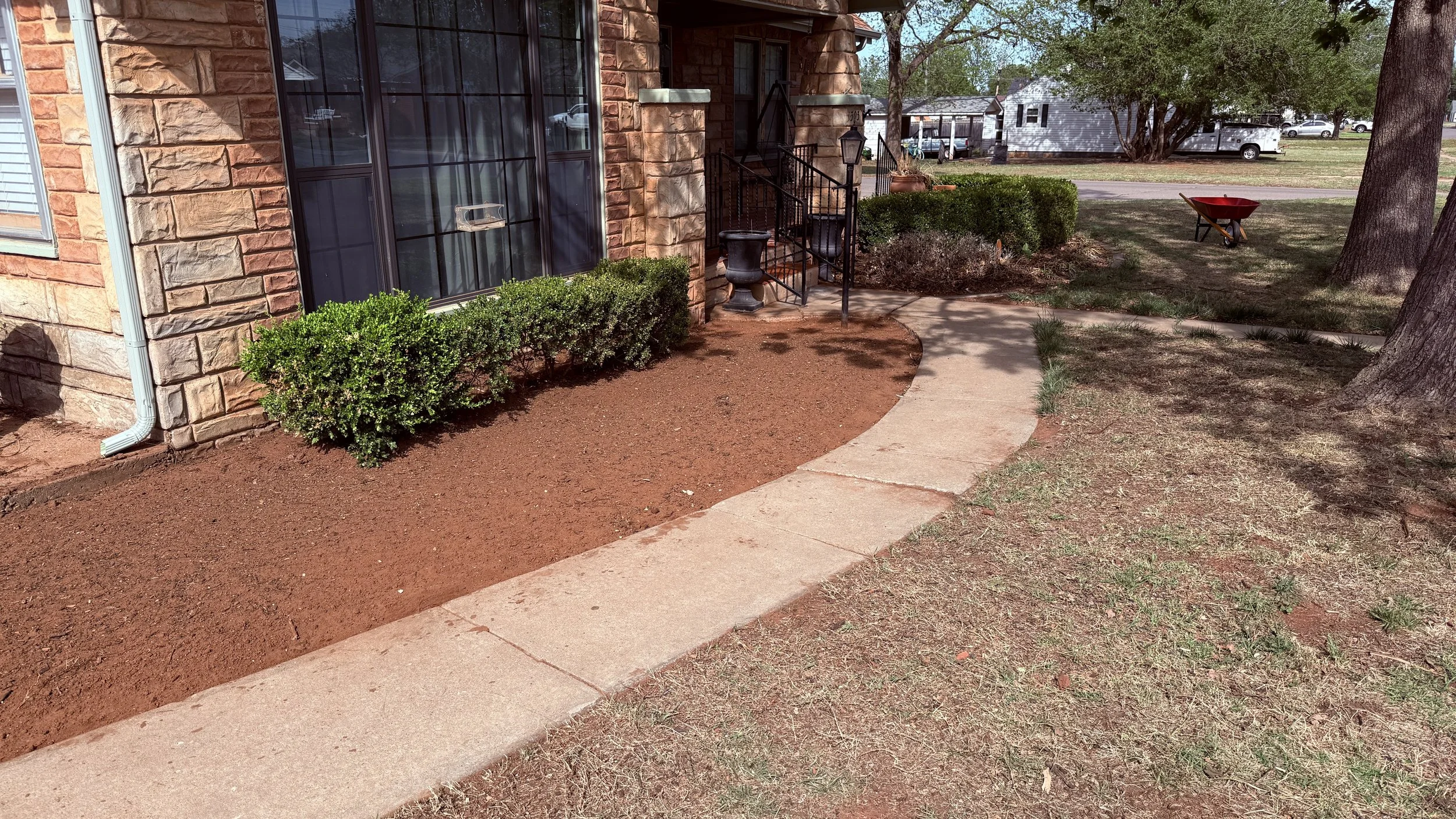 Front of a house with large front window, stone exterior, steps with black railings, small bush, dirt flower bed, concrete walkway, trees, and wheelbarrow in yard.