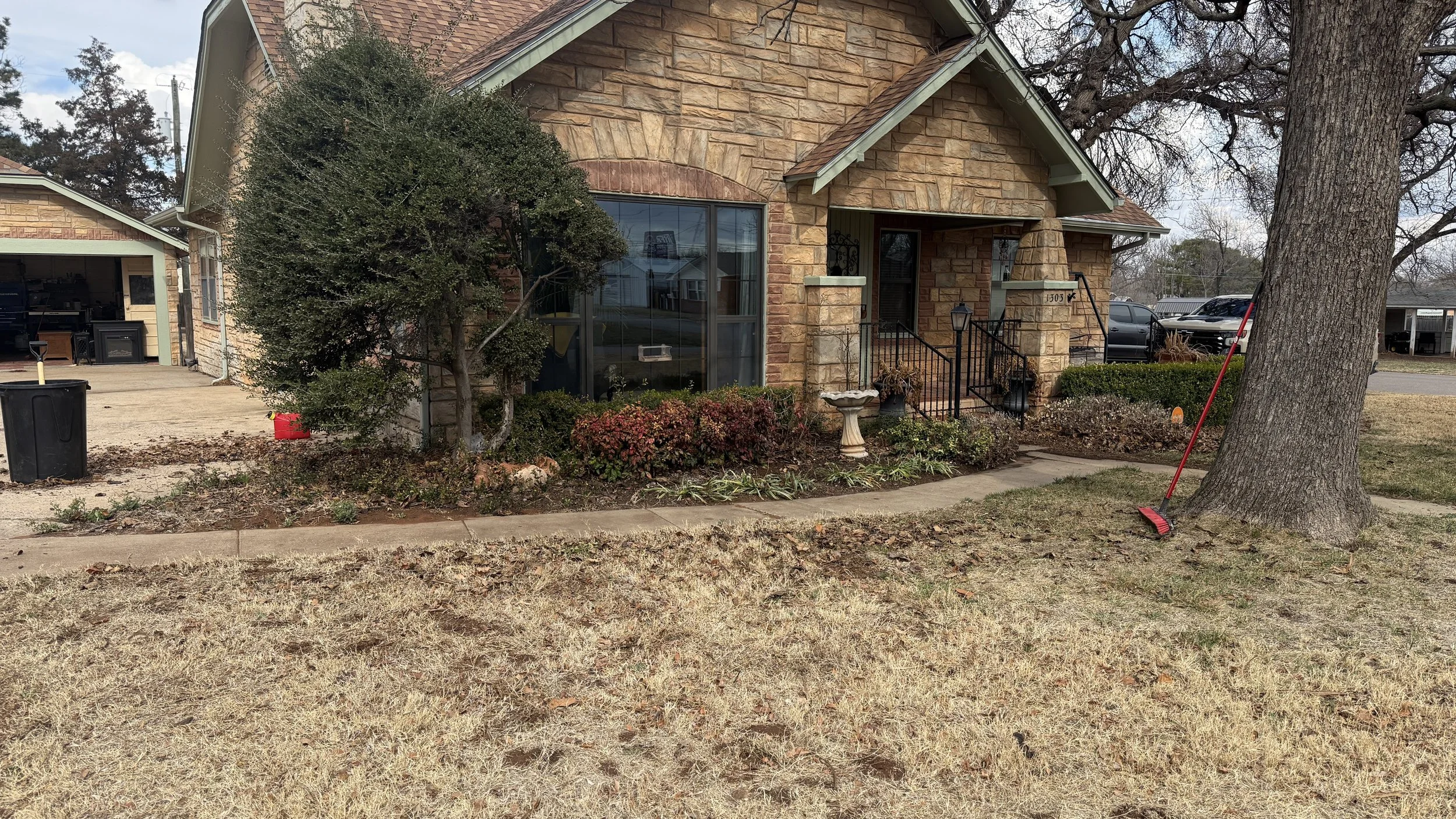 A small brick house with a brown shingled roof, front porch with black railing, large window, and garden bed with bushes and flowers. A large tree leans over the front yard, with a red broom leaning against it. The lawn appears dry and patchy, and there are parked cars and other houses in the background.