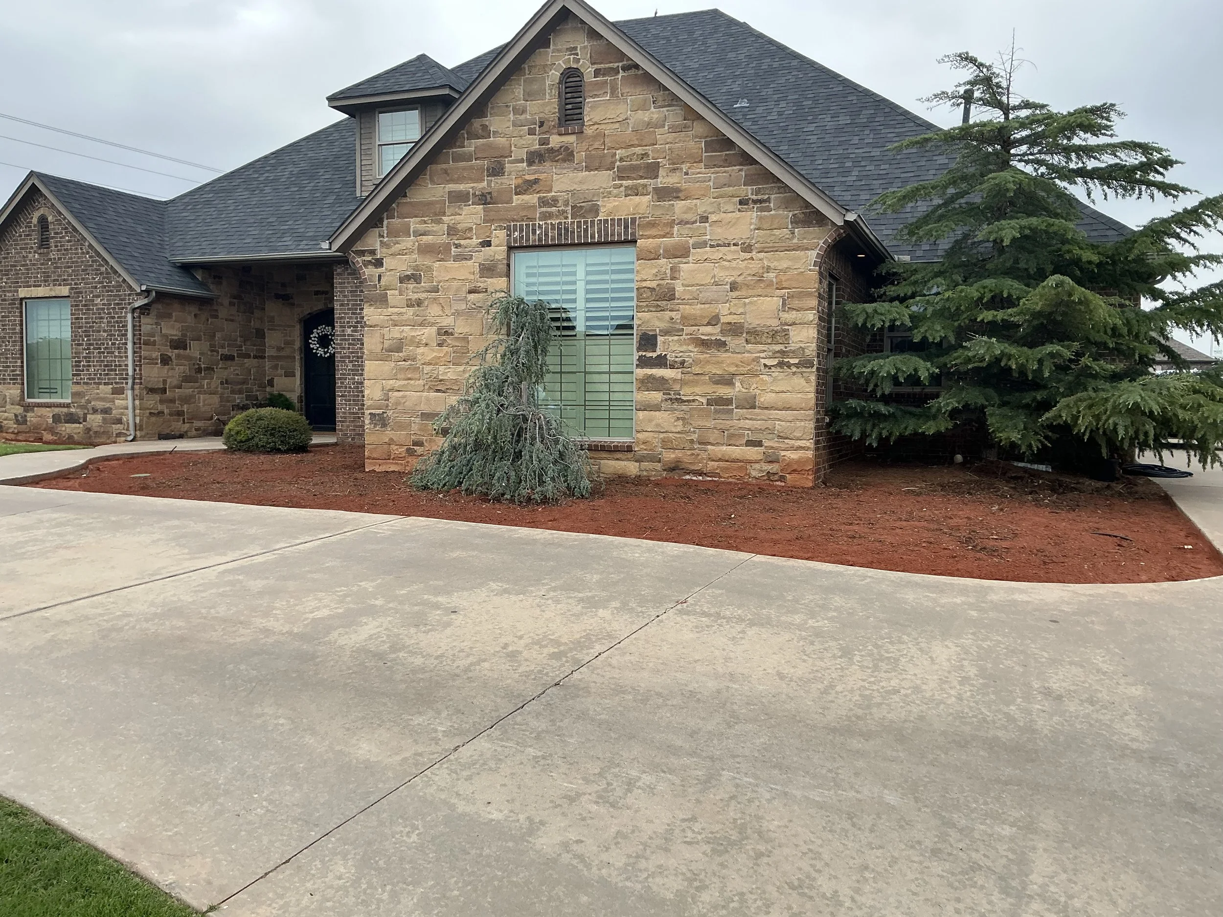 Front view of a house with a stone and brick exterior, large windows with white blinds, and surrounded by a manicured yard with a small evergreen tree and bushes. The driveway is concrete.
