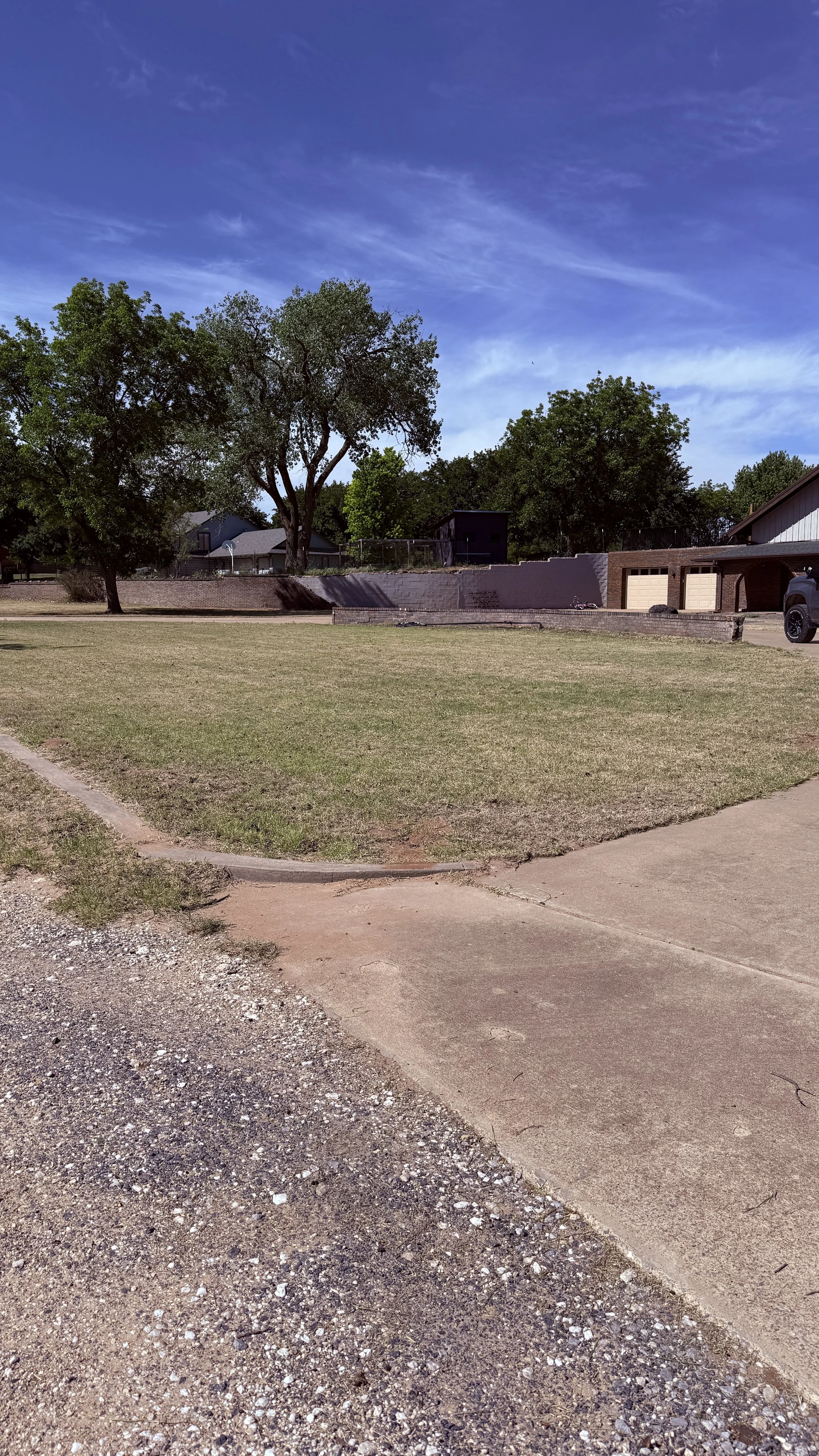 A residential backyard with a large grassy area, a few large trees, a brick retaining wall, and a two-car garage attached to a house. The sky is partly cloudy with blue and white wispy clouds.