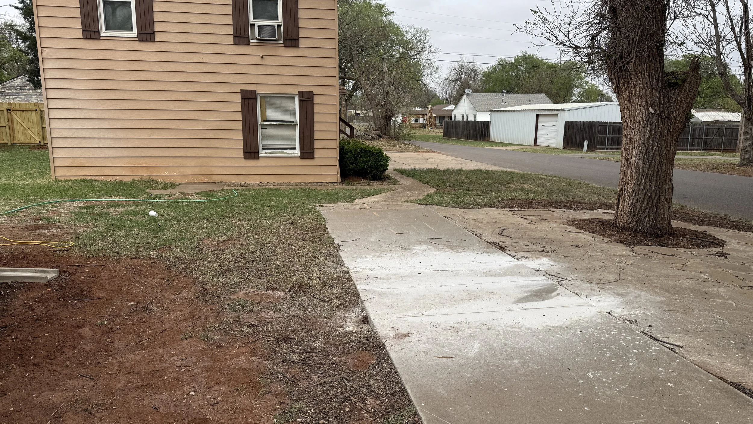 A backyard with a house on the left, showing window shutters, a sidewalk, a large tree, a driveway, and a white garage in the background.