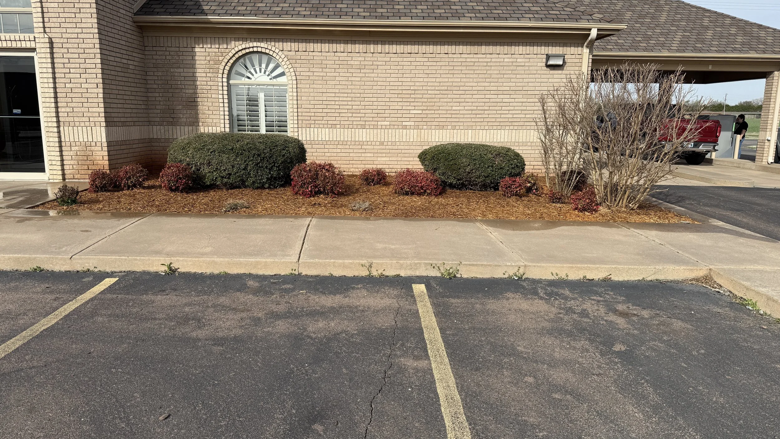 A brick building with a window and outdoor bushes, parking lot in the foreground with yellow parking lines, some shrubs and leafless trees near the building, and a person near a red truck in the background.