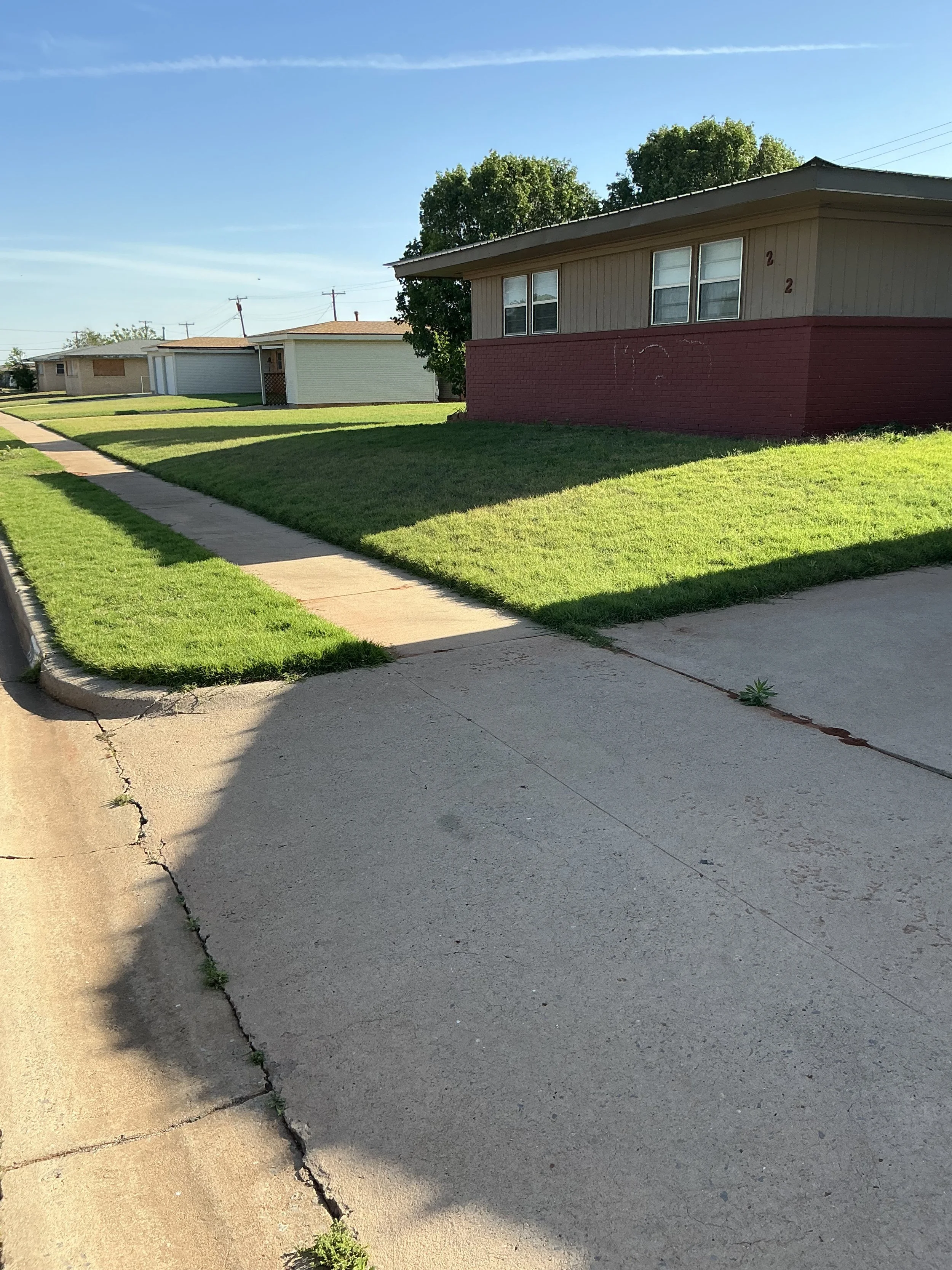 A sidewalk alongside a grassy lawn with a house in the background on a clear sunny day.