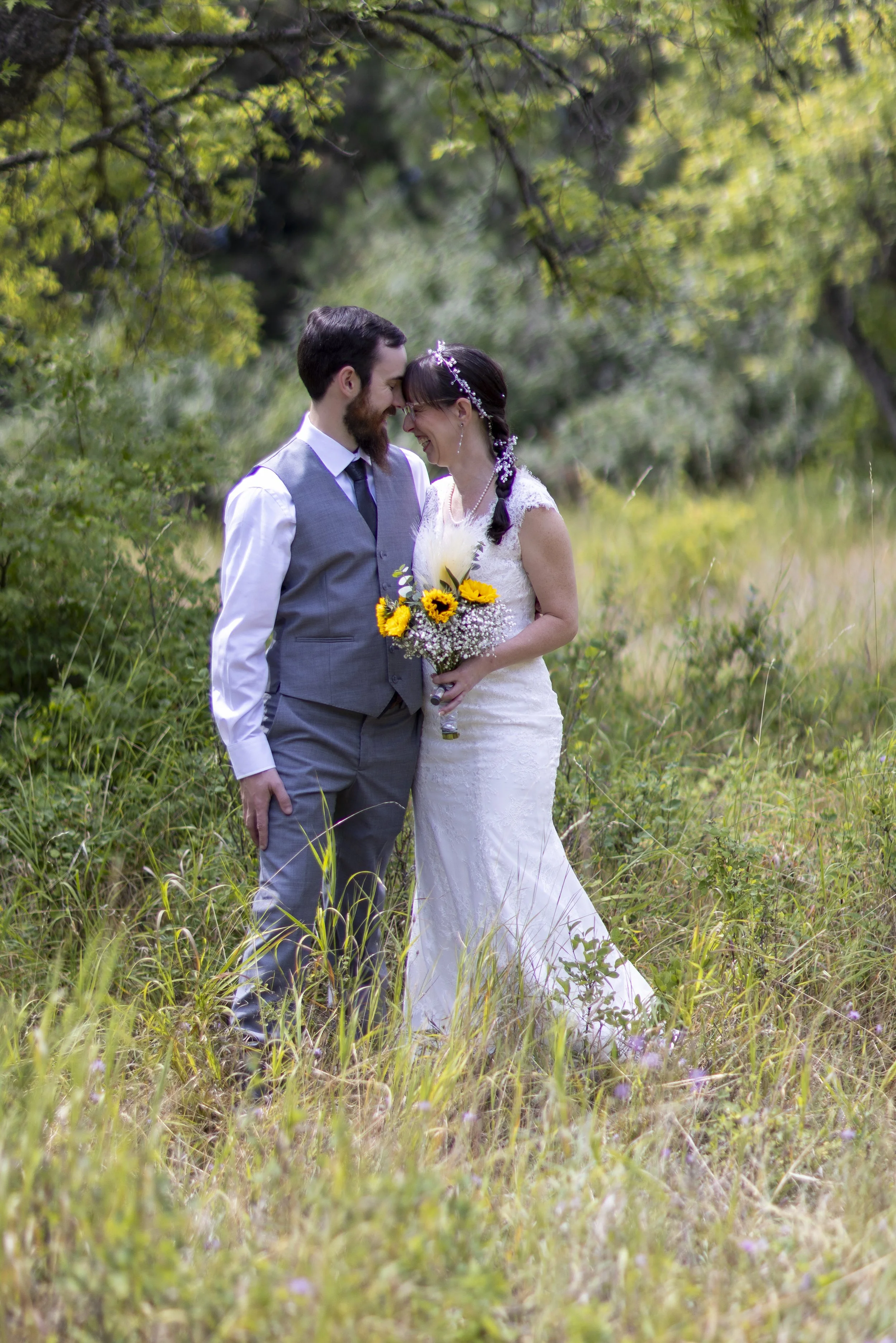 A couple in wedding attire standing close together in a grassy outdoor setting, smiling and touching foreheads, with the bride holding a bouquet of sunflowers and other flowers, surrounded by lush green trees.
