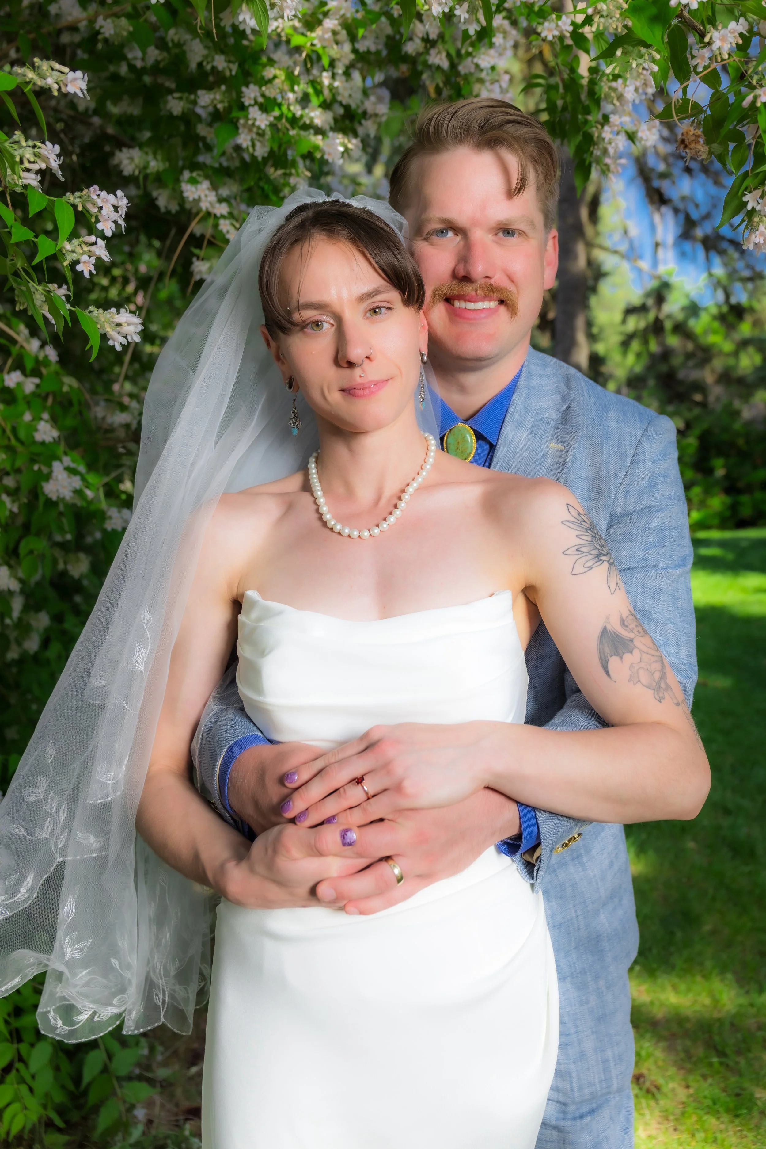 A woman in a wedding dress with a veil and pearl necklace, and a man in a blue suit, posing outdoors among flowering trees.