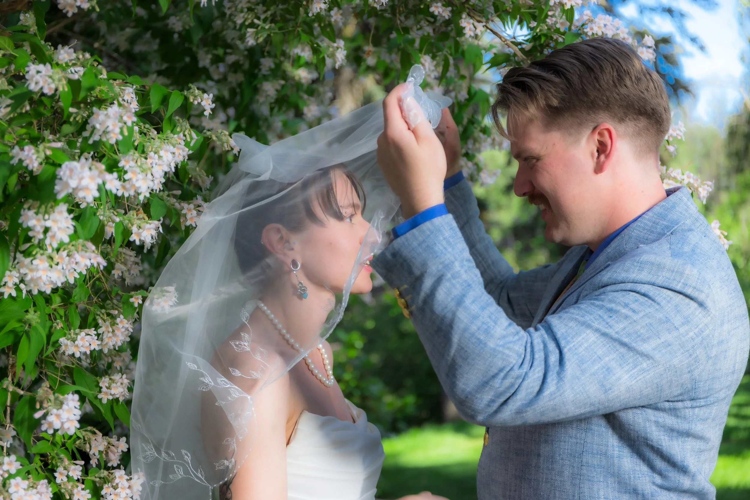 A couple participates in a wedding ceremony outdoors among blossoming trees. The bride is wearing a white dress, pearl jewelry, and a wedding veil. The groom is in a light blue suit. They are smiling, and the groom is lifting a sheer veil over the br