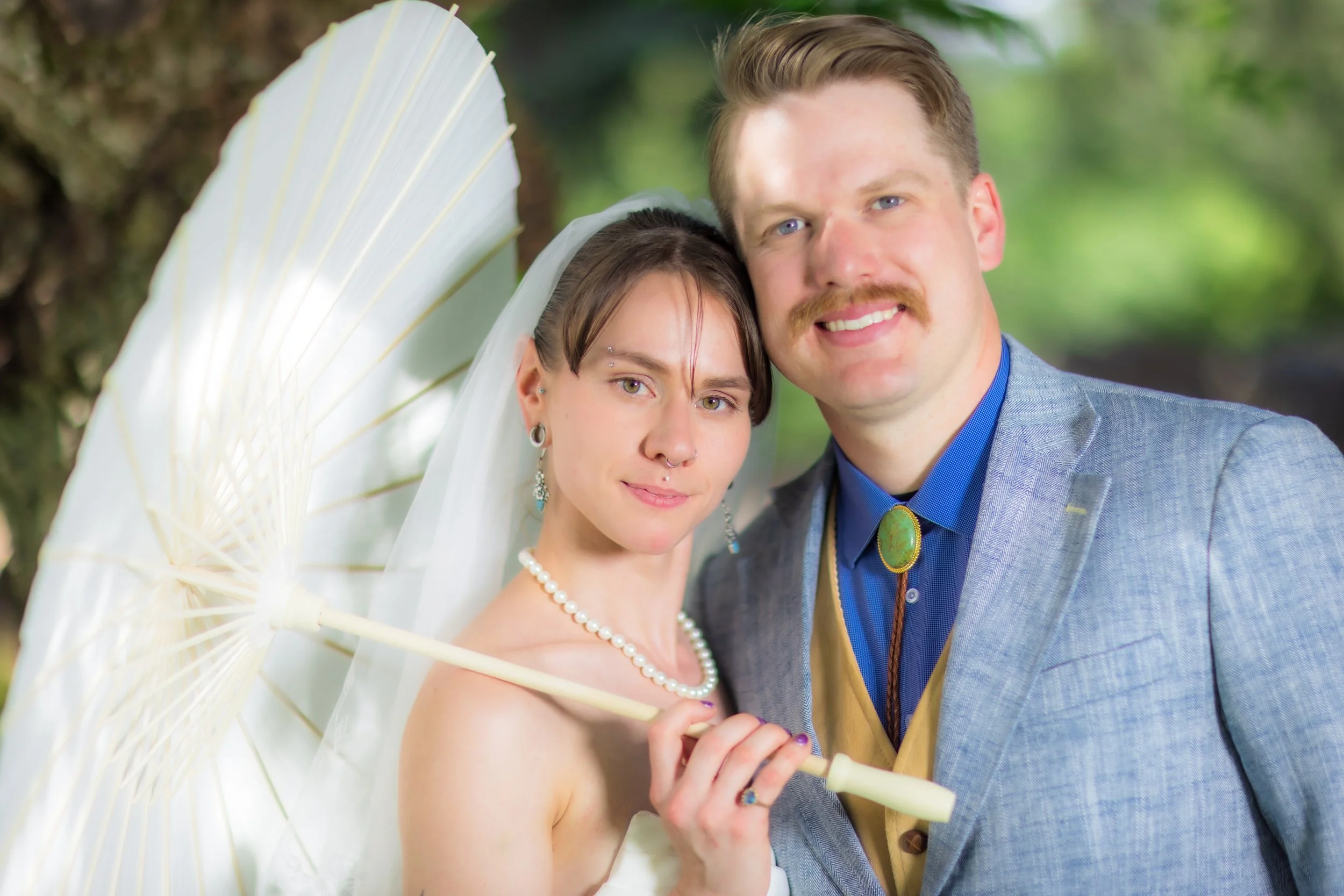 A bride and groom taking a selfie outdoors, with the bride holding a white parasol, both smiling, surrounded by greenery.