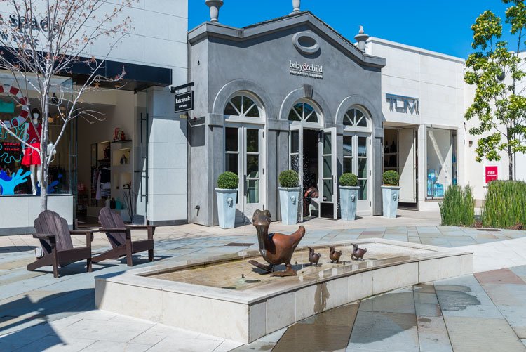 Outdoor shopping area with a duck sculpture fountain, storefronts, and seating