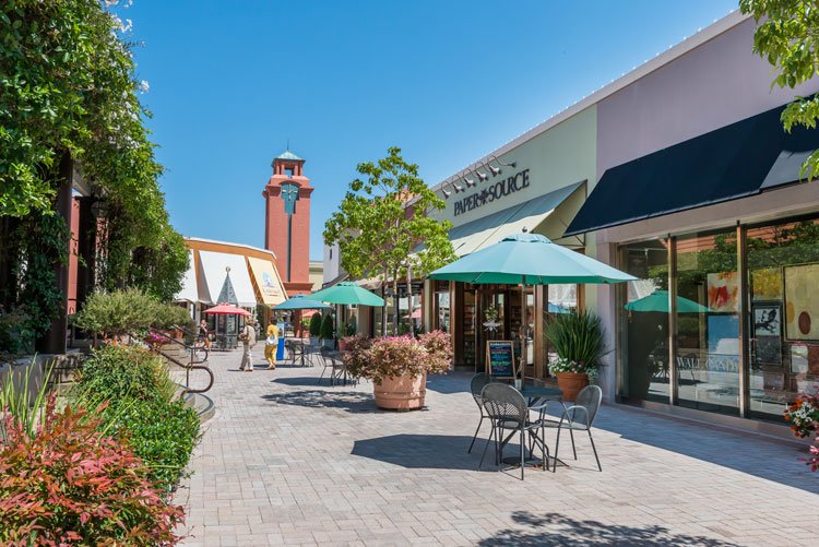Outdoor shopping center with store Paper Source, patio seating, green umbrellas, brick pathway, trees and plants, red clock tower in background.