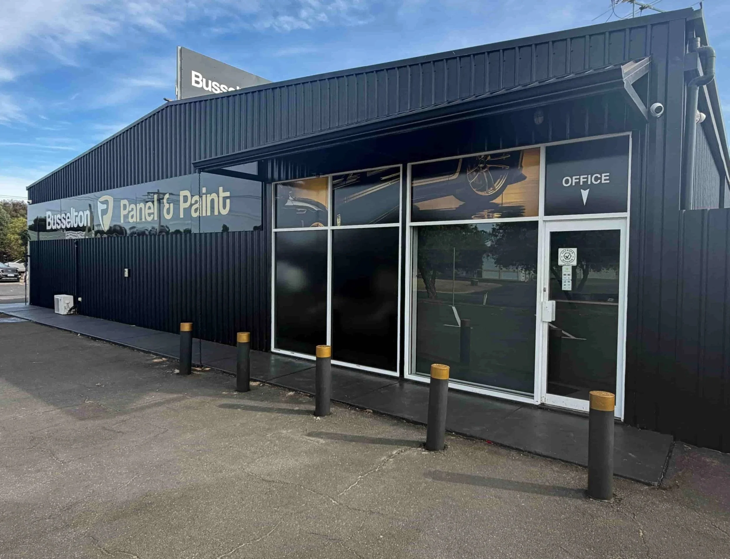 Exterior of a black commercial building with a sign that reads 'Busselton Panel & Paint,' large glass windows, and an entry door labeled 'Office' with a directional arrow.