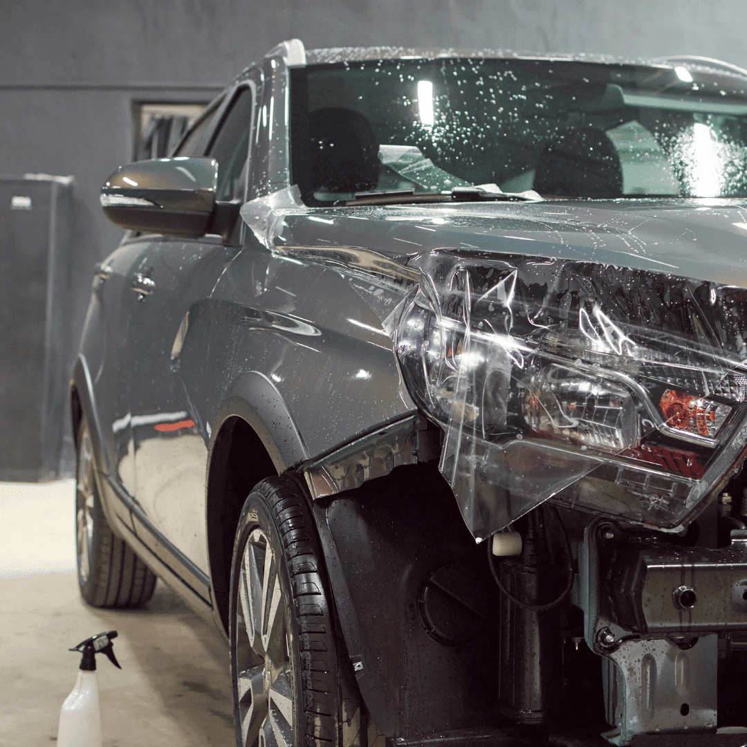Silver SUV with front-end damage and a clear plastic protective sheet over the damaged area, parked indoors with cleaning or repair supplies nearby.