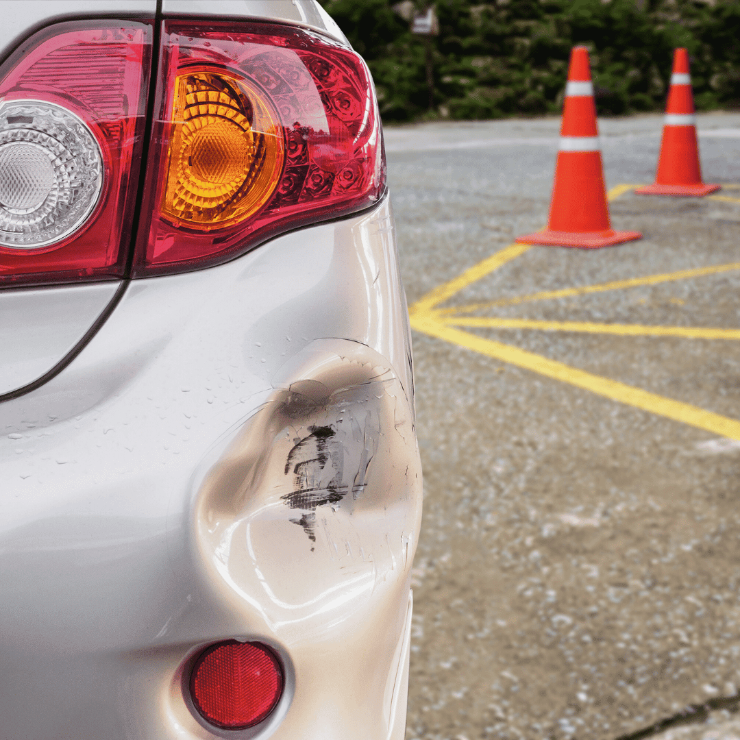 Damaged rear left side of a silver car with a dent and scratches near the taillight, parked in a parking lot with orange cones in the background.