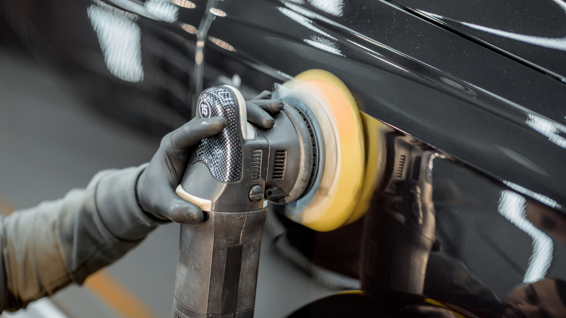 A person wearing black gloves polishing a shiny black car with a yellow buffer machine.