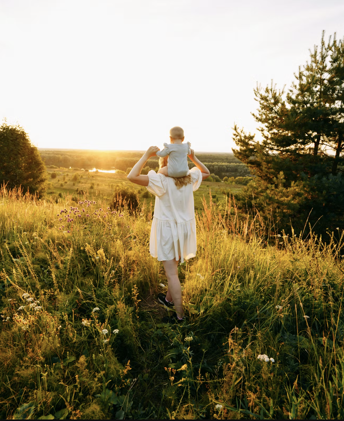 A woman walking through a grassy field with a child on her shoulders at sunset.