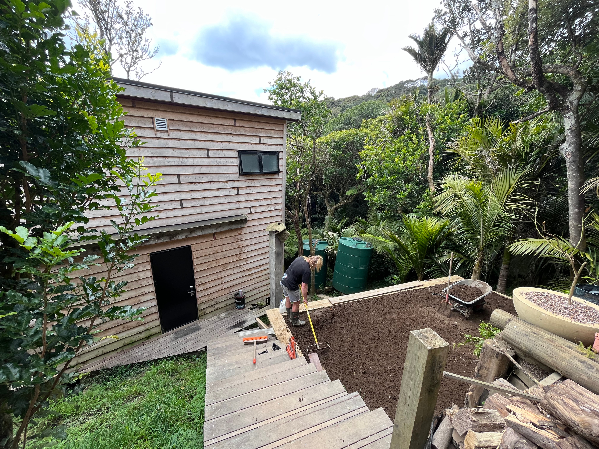 Person gardening in a backyard with a wooden shed, trees, and various gardening tools and supplies.