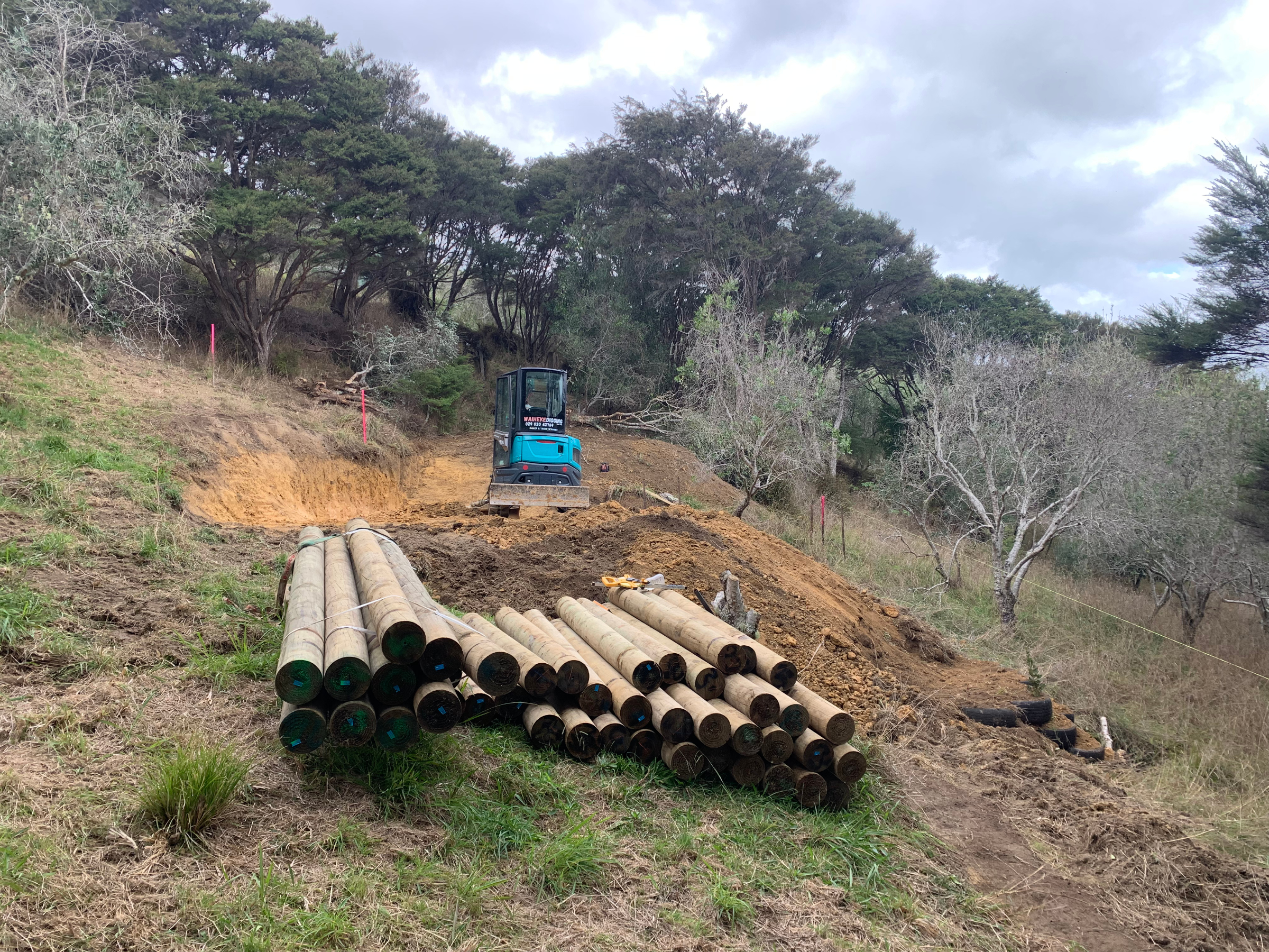 Construction site on a hillside with a small excavator and several stacked wooden pipes.