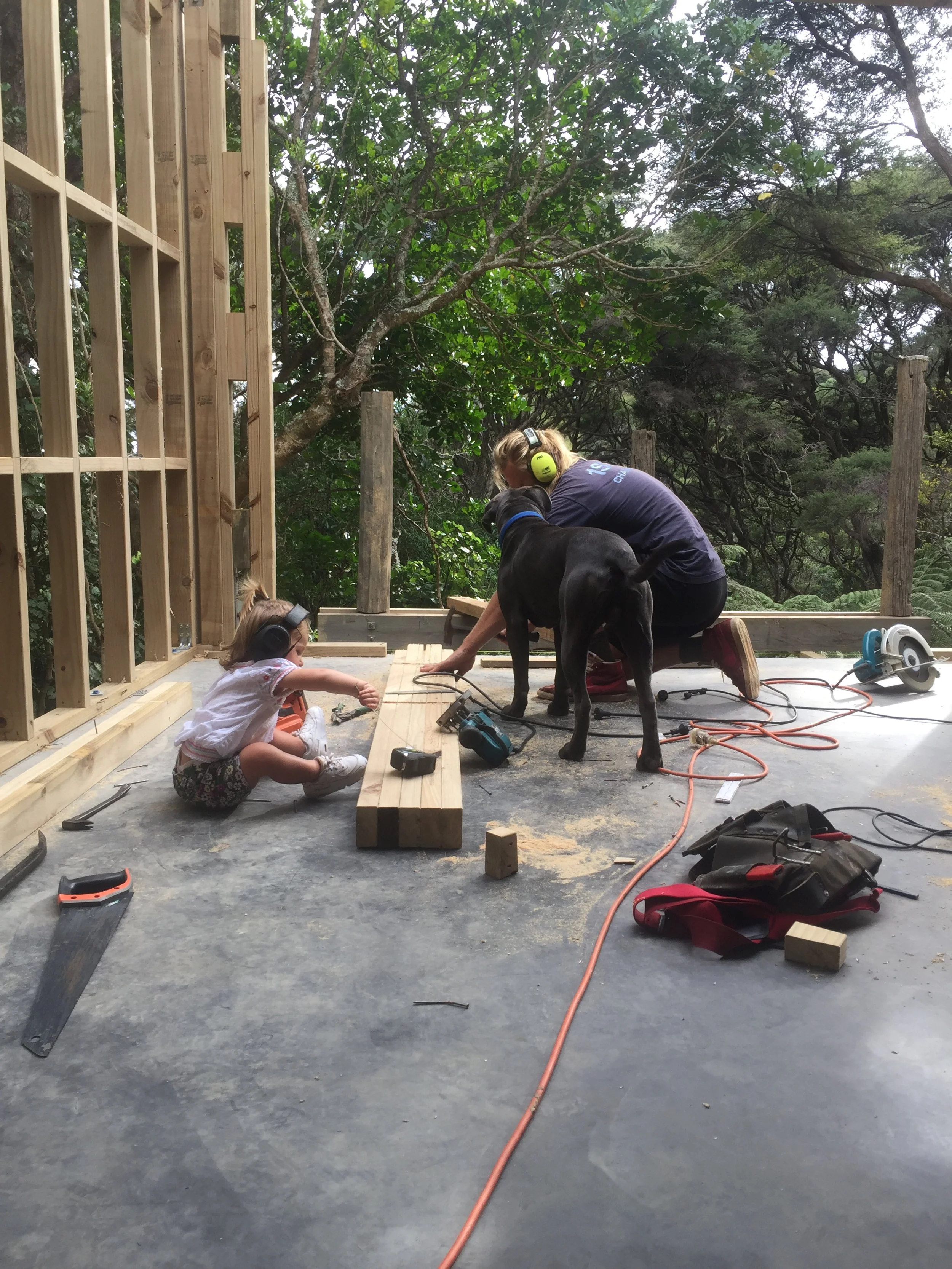 A woman, a young girl, and a dog working on a construction project outdoors. The woman and girl are wearing ear protection, and the woman is using power tools while the girl watches. There are wooden beams, tools, and electrical cords on the floor, with trees in the background.