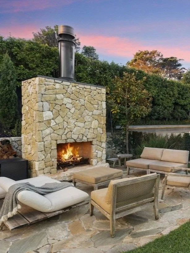 An outdoor living area with a stone fireplace burning with a fire, surrounded by comfortable chairs, coffee table, and a firewood stack, with greenery and a sunset sky in the background.