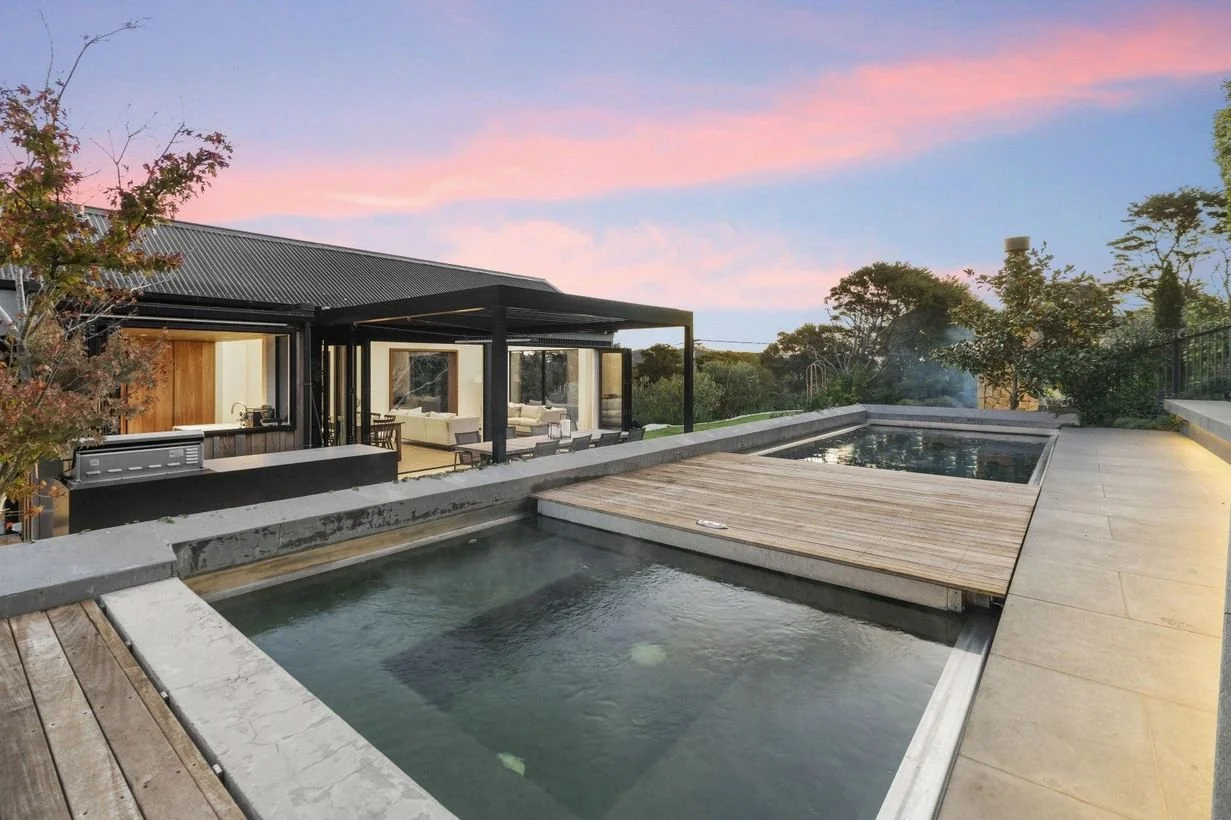 Modern house with outdoor pool area at sunset, surrounded by trees and a pink and blue sky.