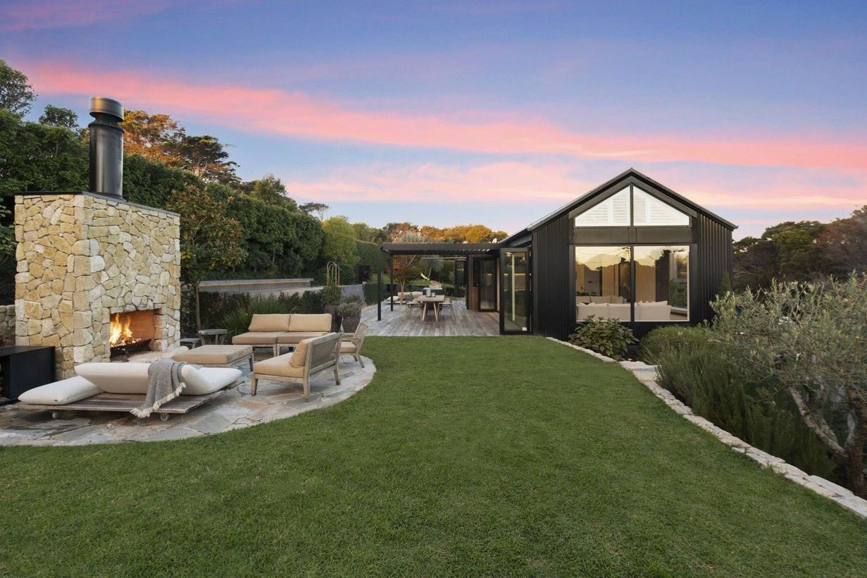 Modern backyard with a black house, outdoor fireplace, cushioned chairs, and a wooden deck at sunset.