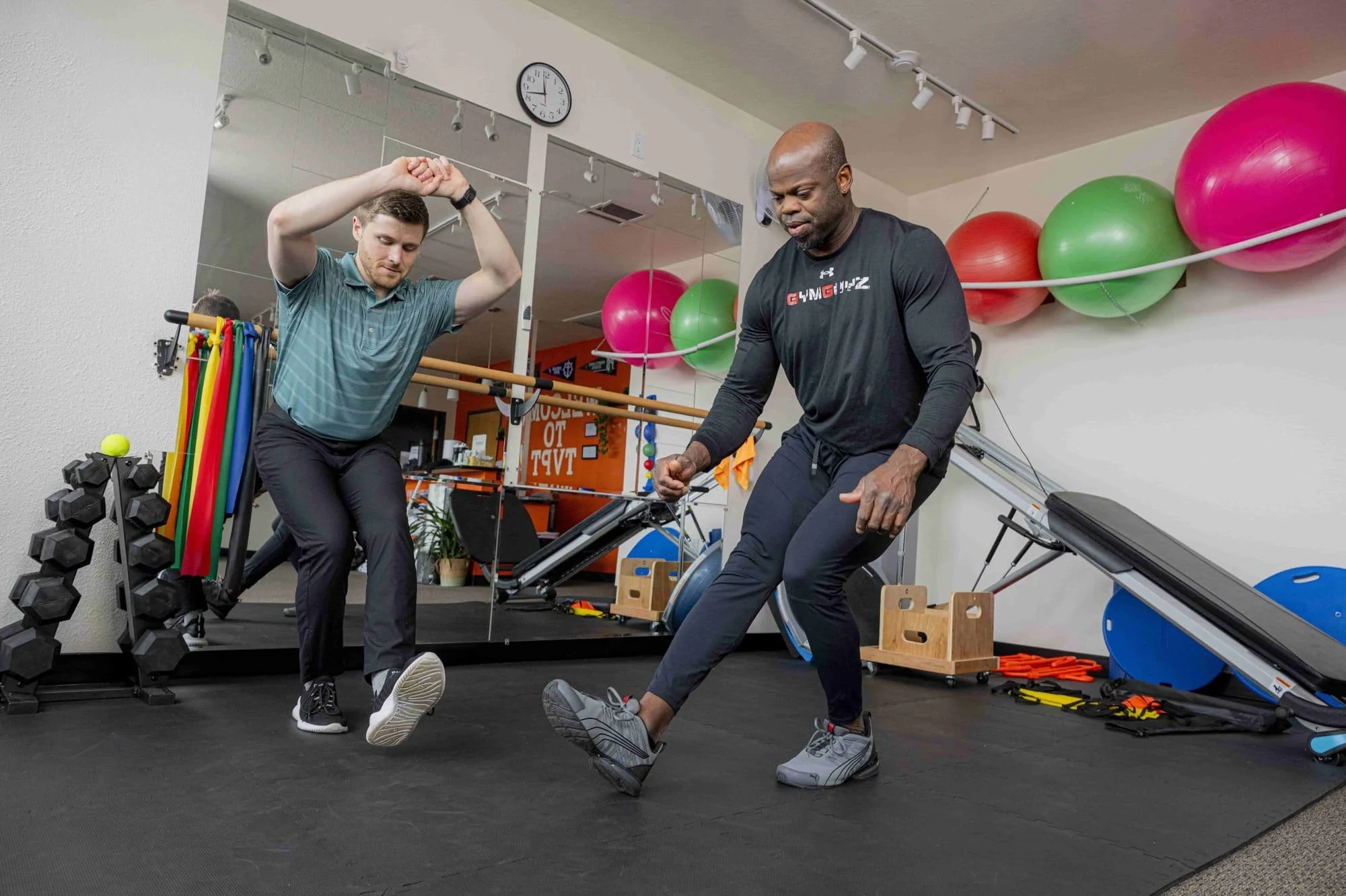 A physical therapist leading a patient through a single-leg squat exercise in a physical therapy clinic's gym area