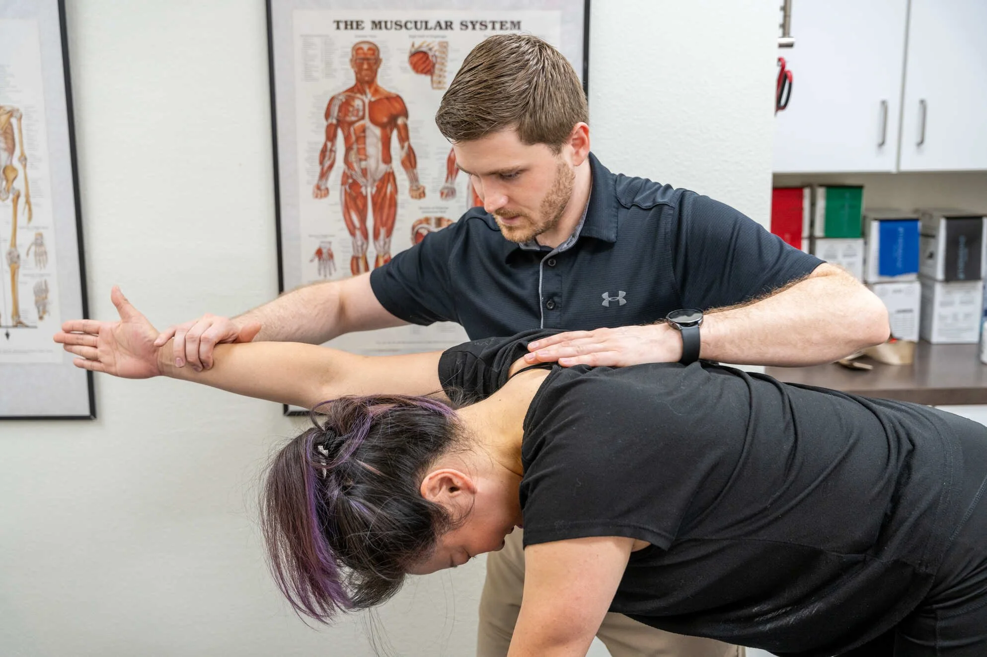 A male physical therapist examining and adjusting the shoulder of a woman who is in a quadruped position performing a scapular stability exercise for shoulder pain. They are in an office with anatomical posters on the wall.