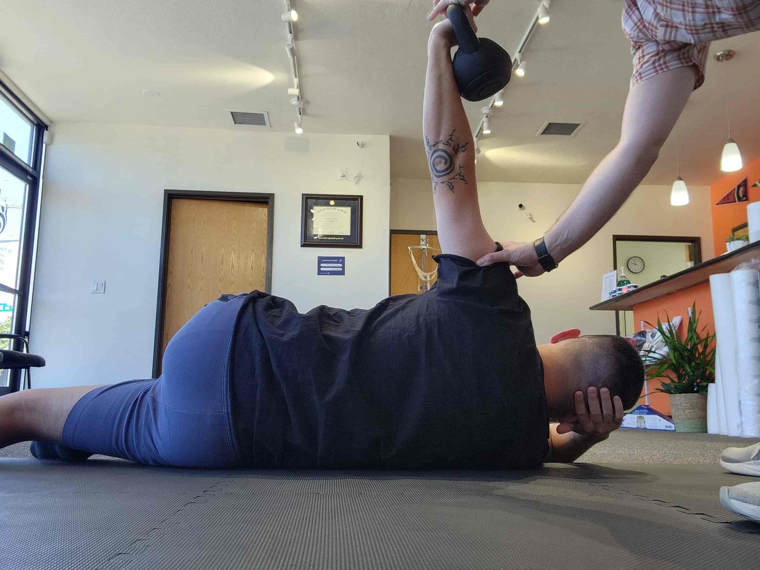 A man lying on the floor performing armbars using a kettle bell with guidance from a PT