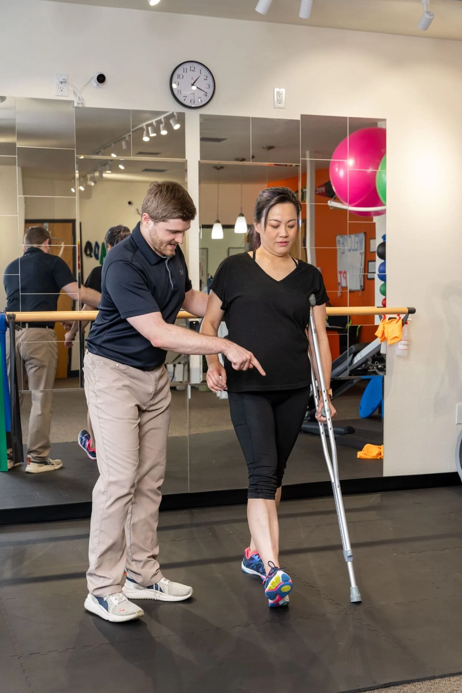 A physical therapist helps a woman walk using crutches in a fitness center with mirror wall reflections and colorful exercise balls.