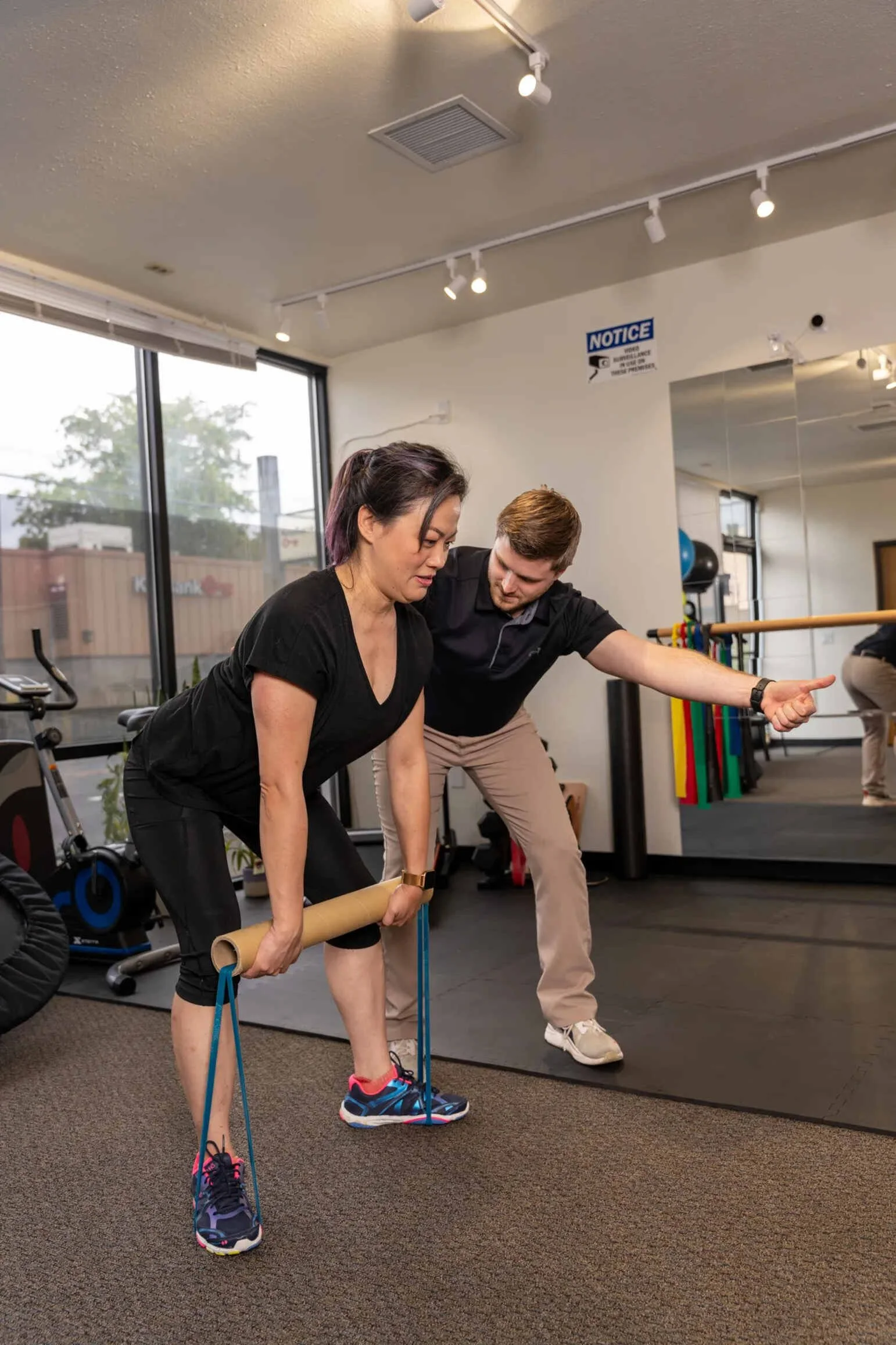 A woman practicing a deadlift exercise with resistance bands, guided by a physical therapist in a PT clinic.