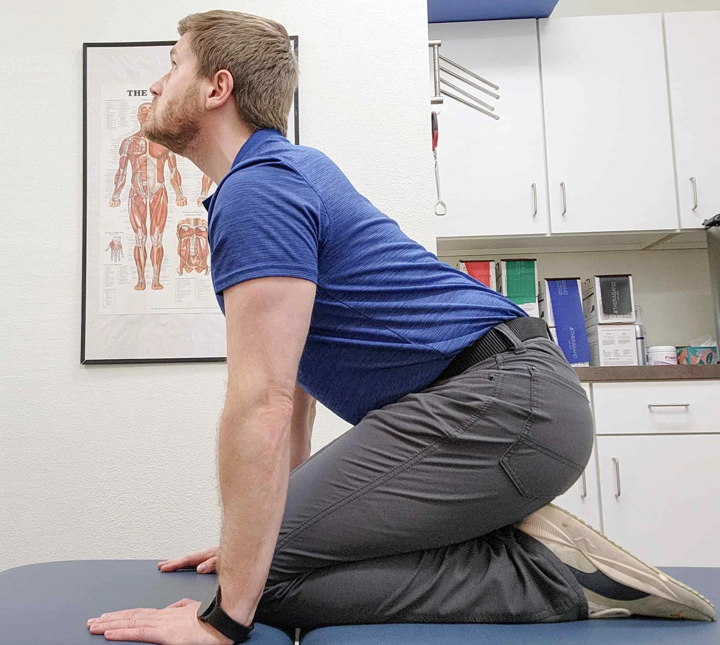 A man in a blue shirt and gray pants performing a stretch on a medical examination table in a clinical setting.