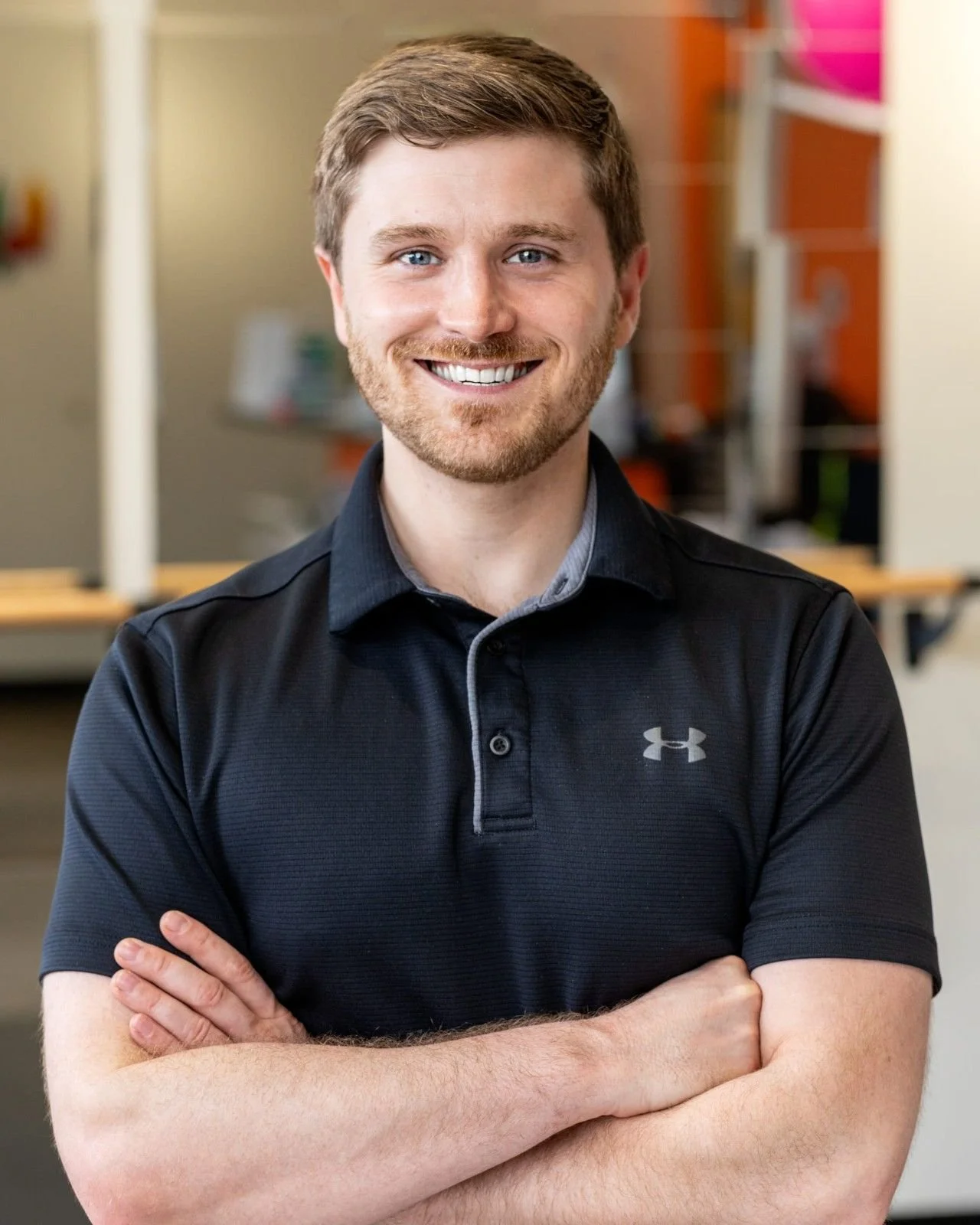 Headshot of a smiling young man with light brown hair and blue eyes wearing a black Under Armour polo shirt, in an indoor setting with colorful background elements posing for a photo.