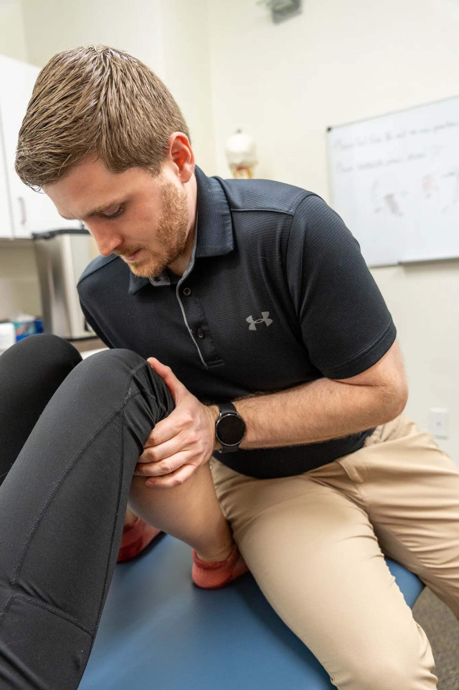 A physiotherapist performs a knee rehabilitation exercise on a patient lying on a treatment table, holding the patient's knee and ankle.