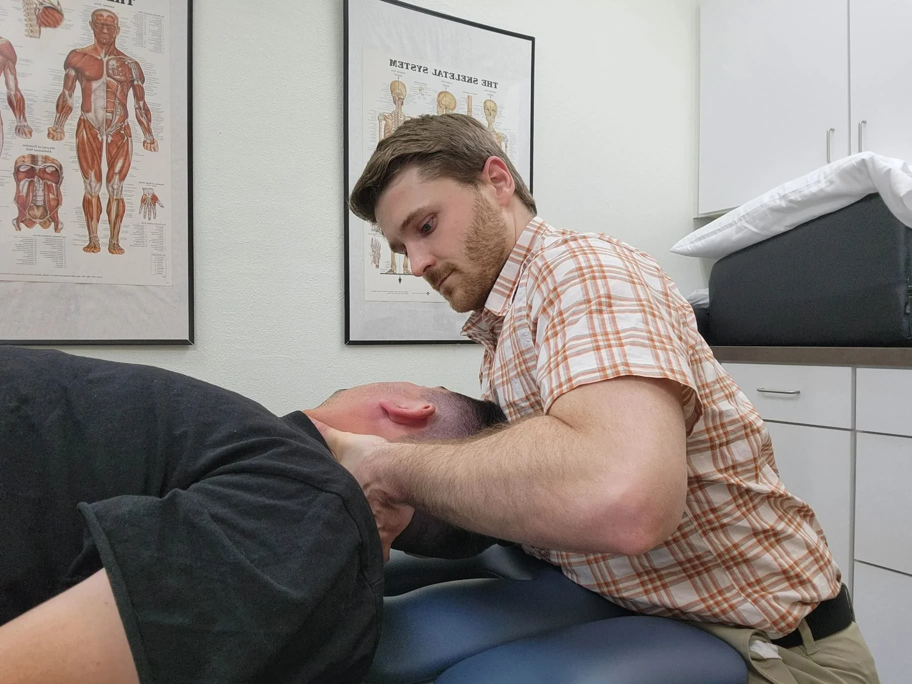 A physical therapist adjusting a patient's neck in an exam room, with anatomical posters on the wall.