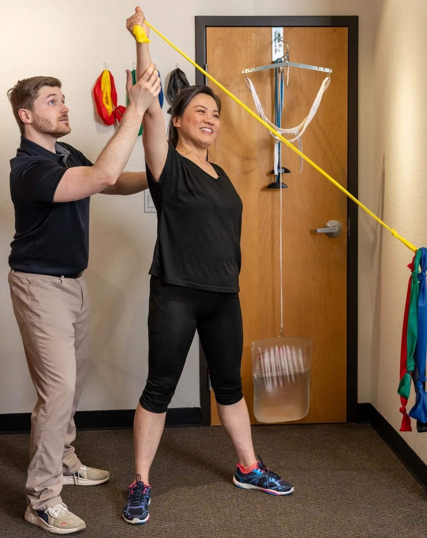 A woman in black workout clothes uses a yellow resistance band, assisted by a physical therapist, to perform upper extremity PNF patterns in a therapy room.