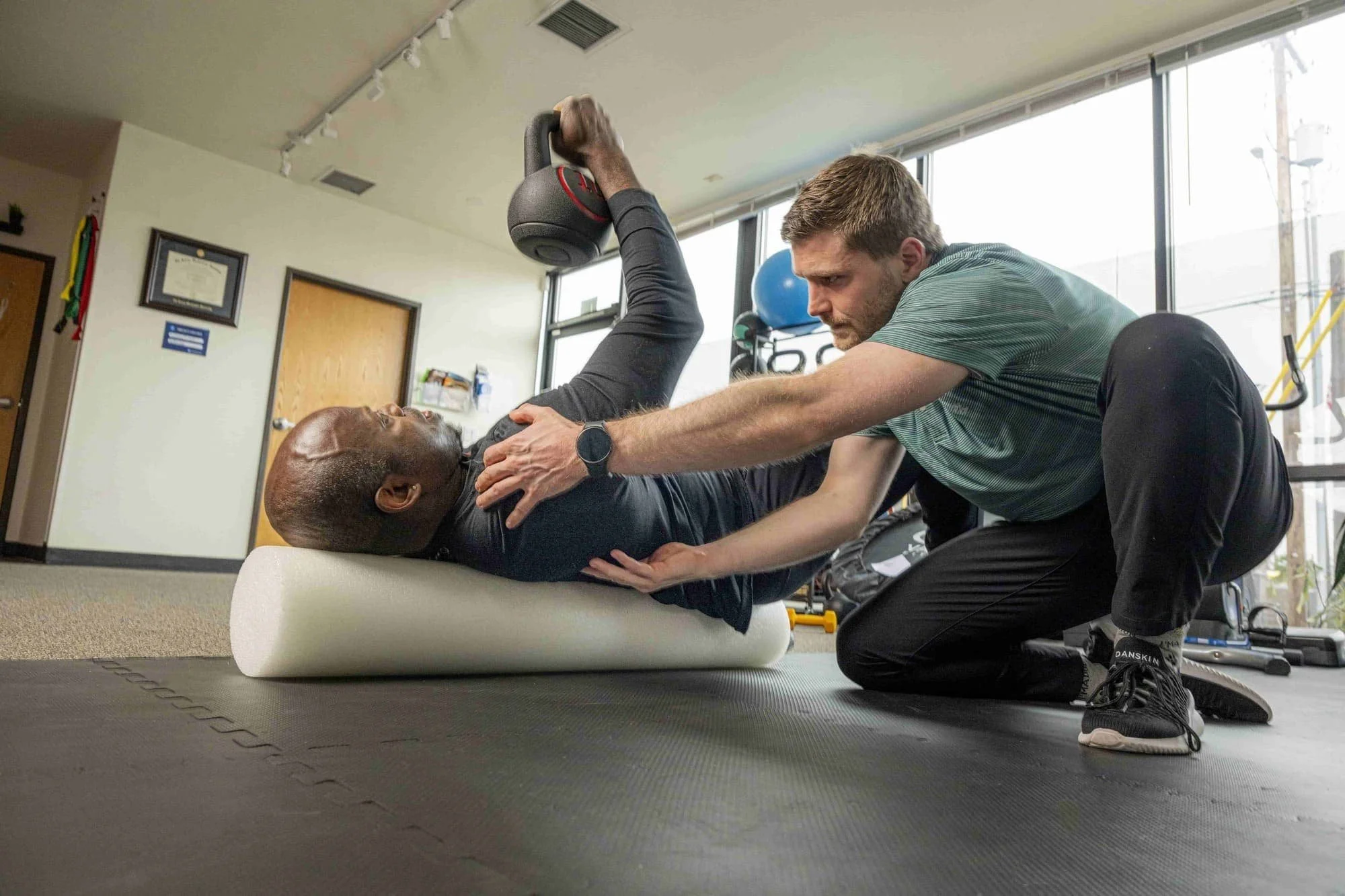 A physical therapist in TVPT's clinic's main floor area is guiding the shoulder blade of a man lying down on a foam roll and pressing a kettlebell upwards like a corkscrew