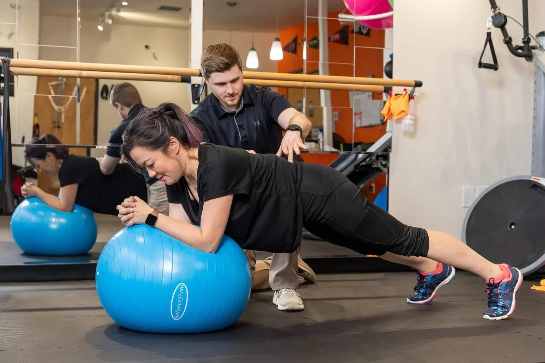 A woman performing a plank on a stability ball in a physical therapy clinic with a physical therapist guiding the exercise.