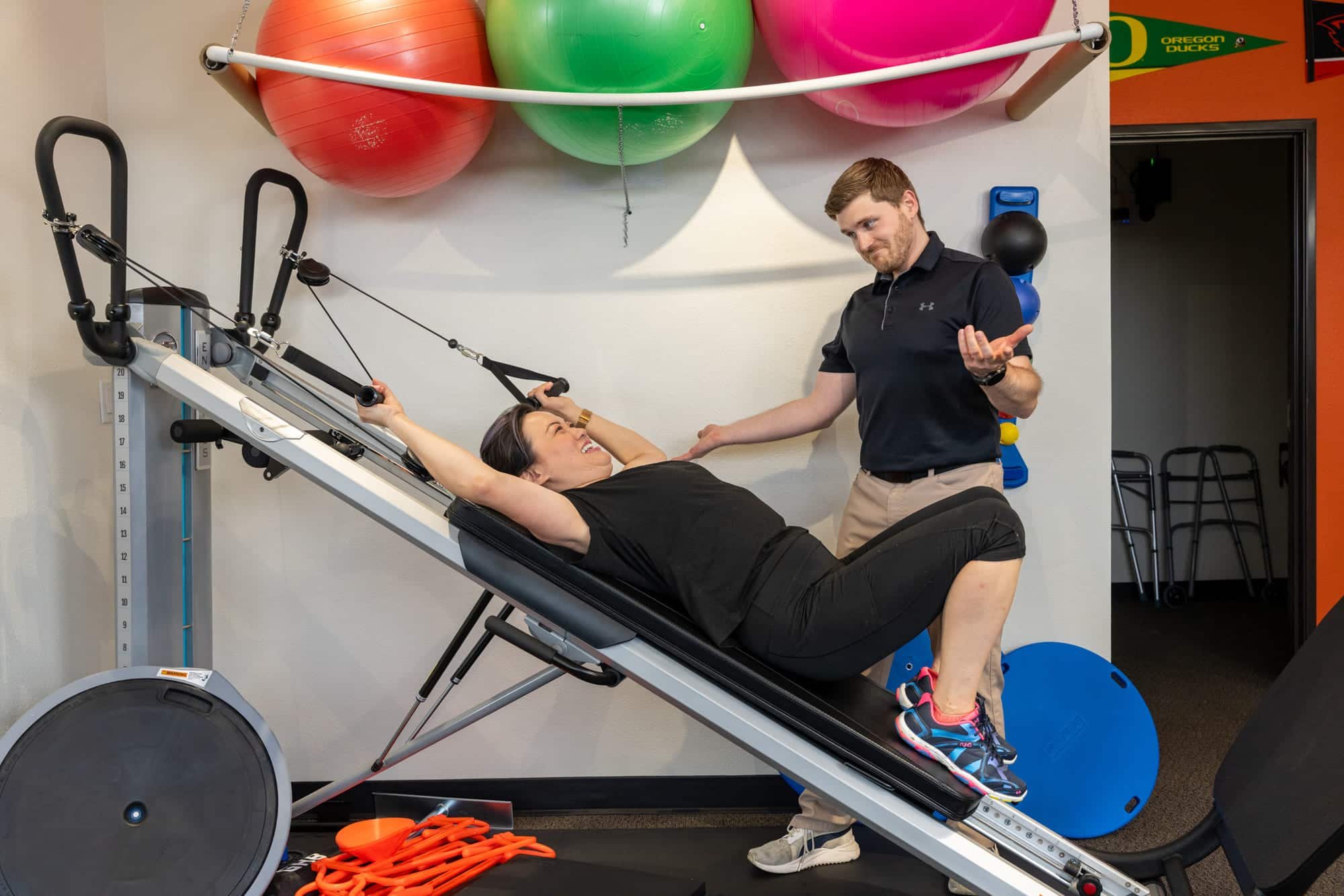 A woman is lying on a slope trainer exercising, with her arms stretched above her head. A physical therapist  beside her is gesturing with his hands, explaining or coaching her during her workout in a PT clinic.