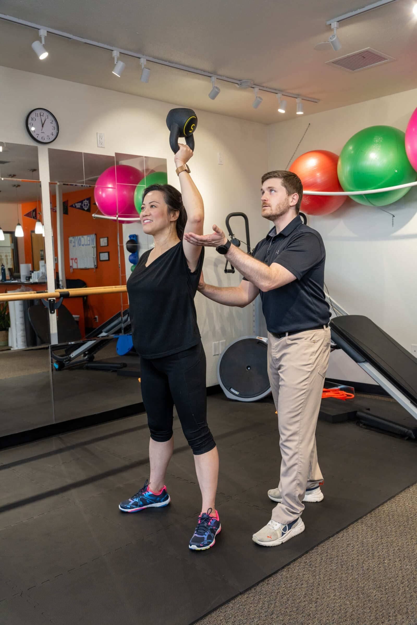 A woman lifting a kettlebell overhead while a physical therapist supervises her in a clinic with colorful exercise balls and mirrors.