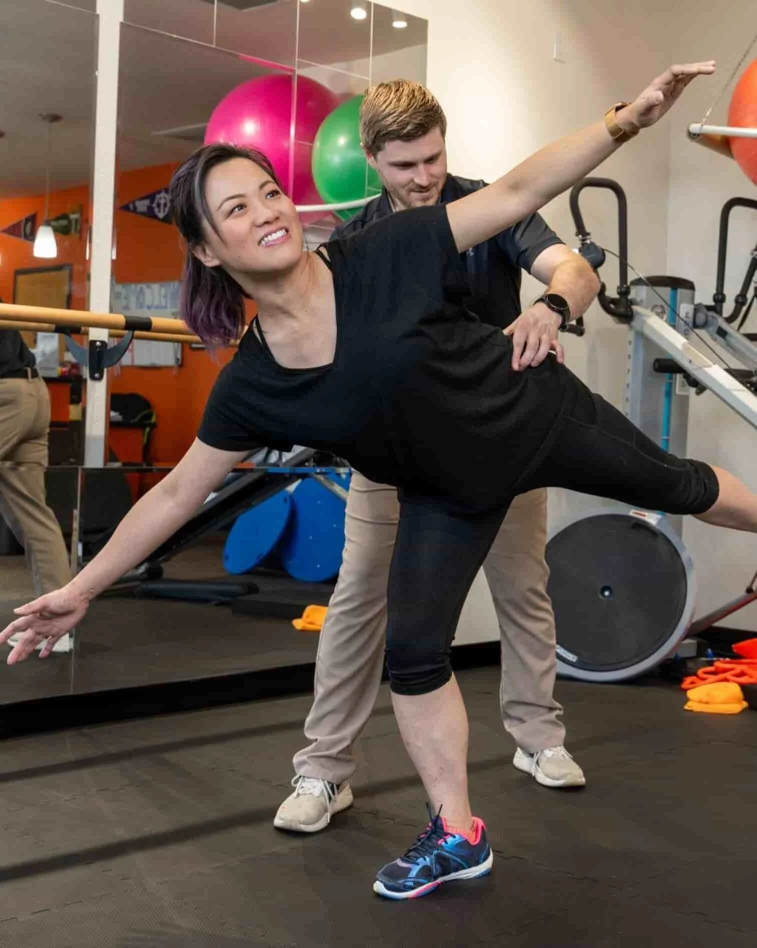 A woman in black athletic wear is performing a balance exercise (hip airplane) with the assistance of a physical therapist in a clinic.