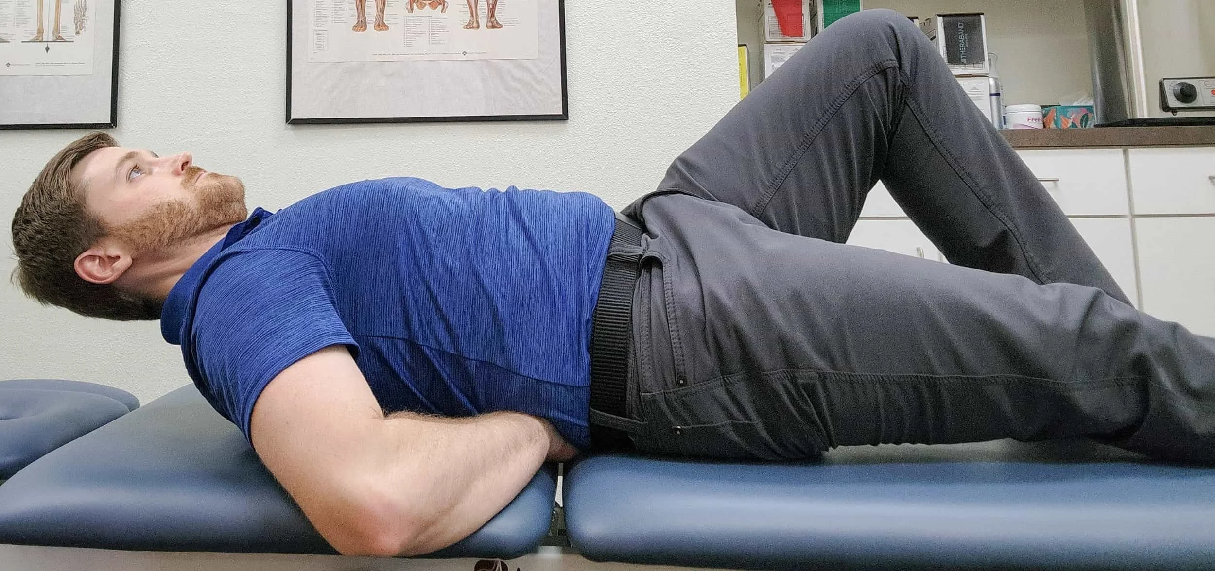 A man lying on a medical examination table performing an ab curl up in a clinical setting with medical posters on the wall.