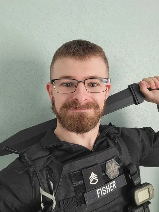A man with glasses and a beard wearing a tactical uniform with the name tag 'Fisher' and holding a large knife over his shoulder.