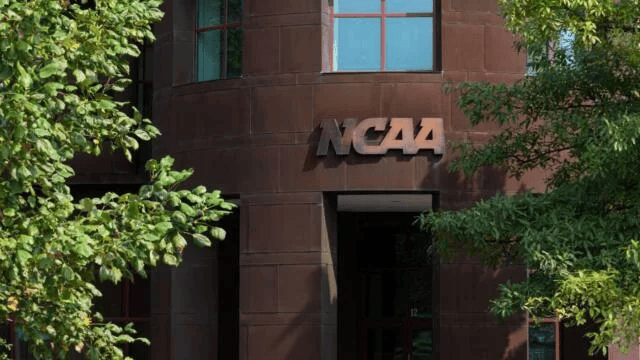 The brown exterior of the NCAA headquarters building is visible through green trees, with the NCAA logo prominently displayed above the entrance.