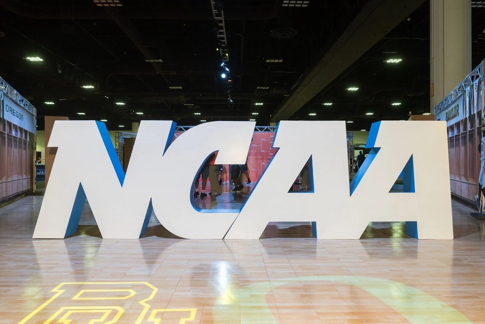 Large, 3D letters of the white NCAA logo with blue trim is displayed on a wooden floor.