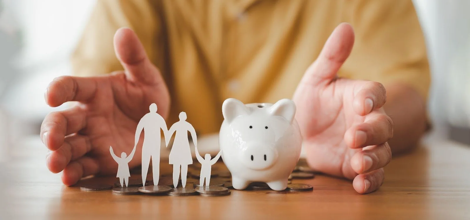 A white piggy bank sits on a pile of coins, flanked by paper cutouts of a family (two adults and two children). A person's hands are cupped protectively around the piggy bank and family.