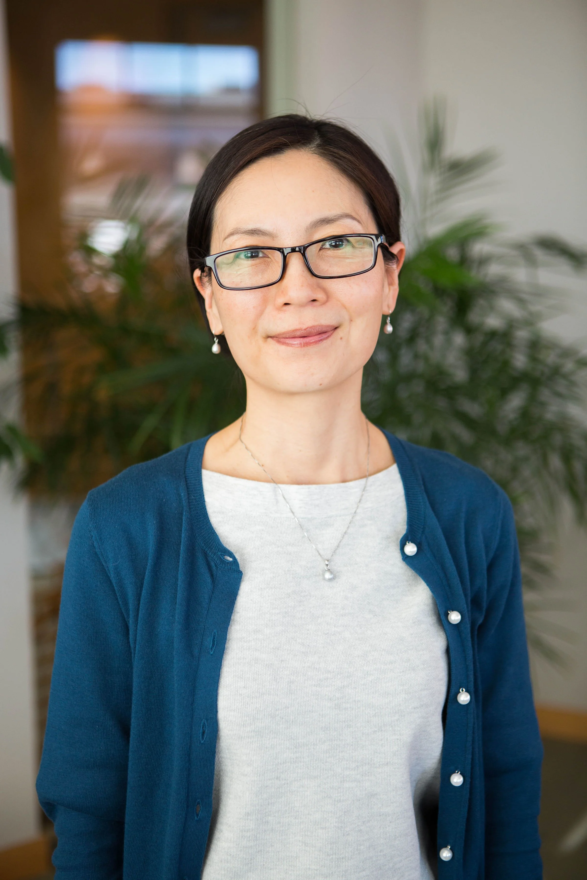 A professional headshot of a woman with dark hair and glasses, wearing a white shirt and blue cardigan, smiling in front of green plants.