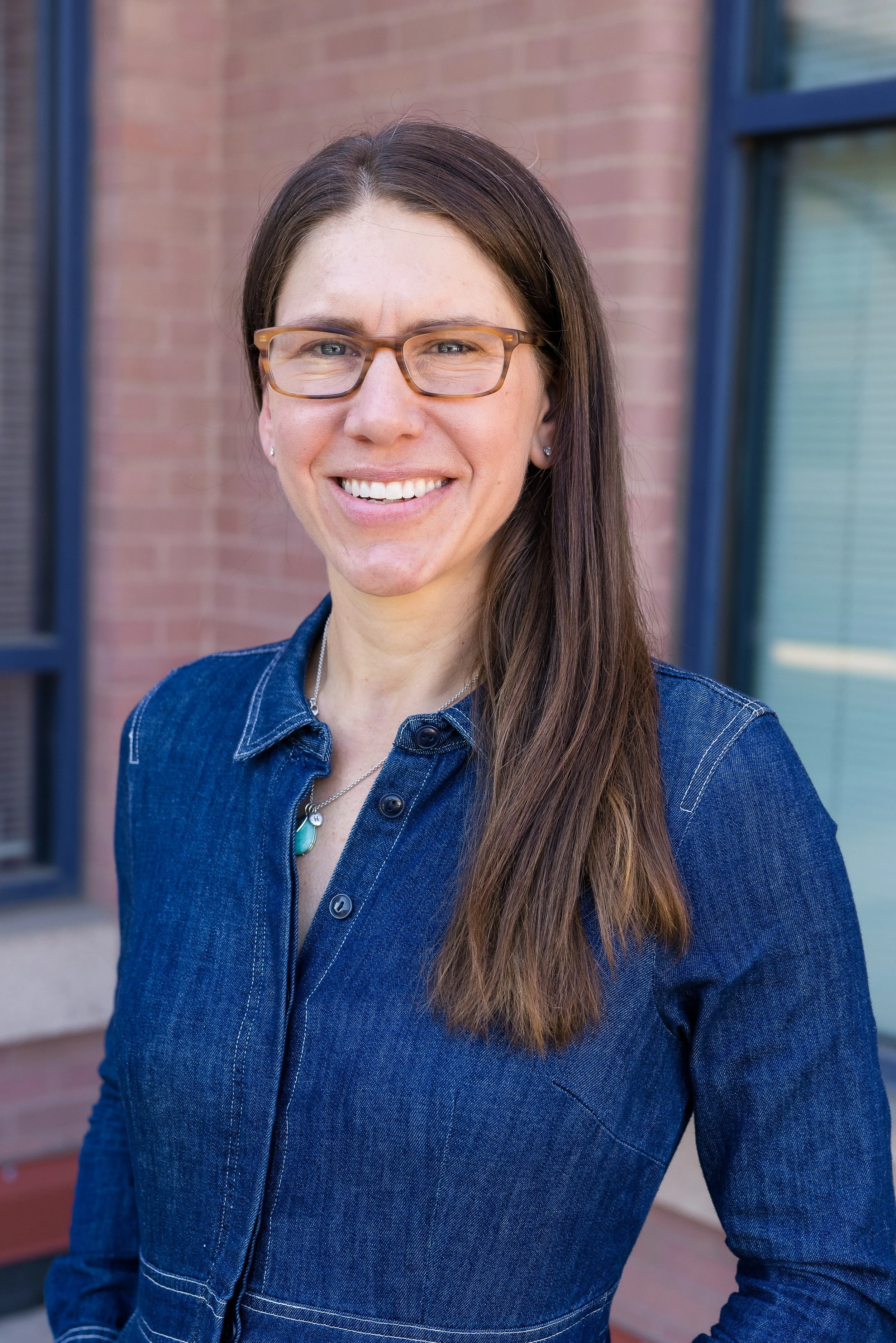 A professional headshot of a smiling woman with long brown hair and glasses, wearing a denim shirt with a turquoise pendant, standing outdoors in front of a brick building.