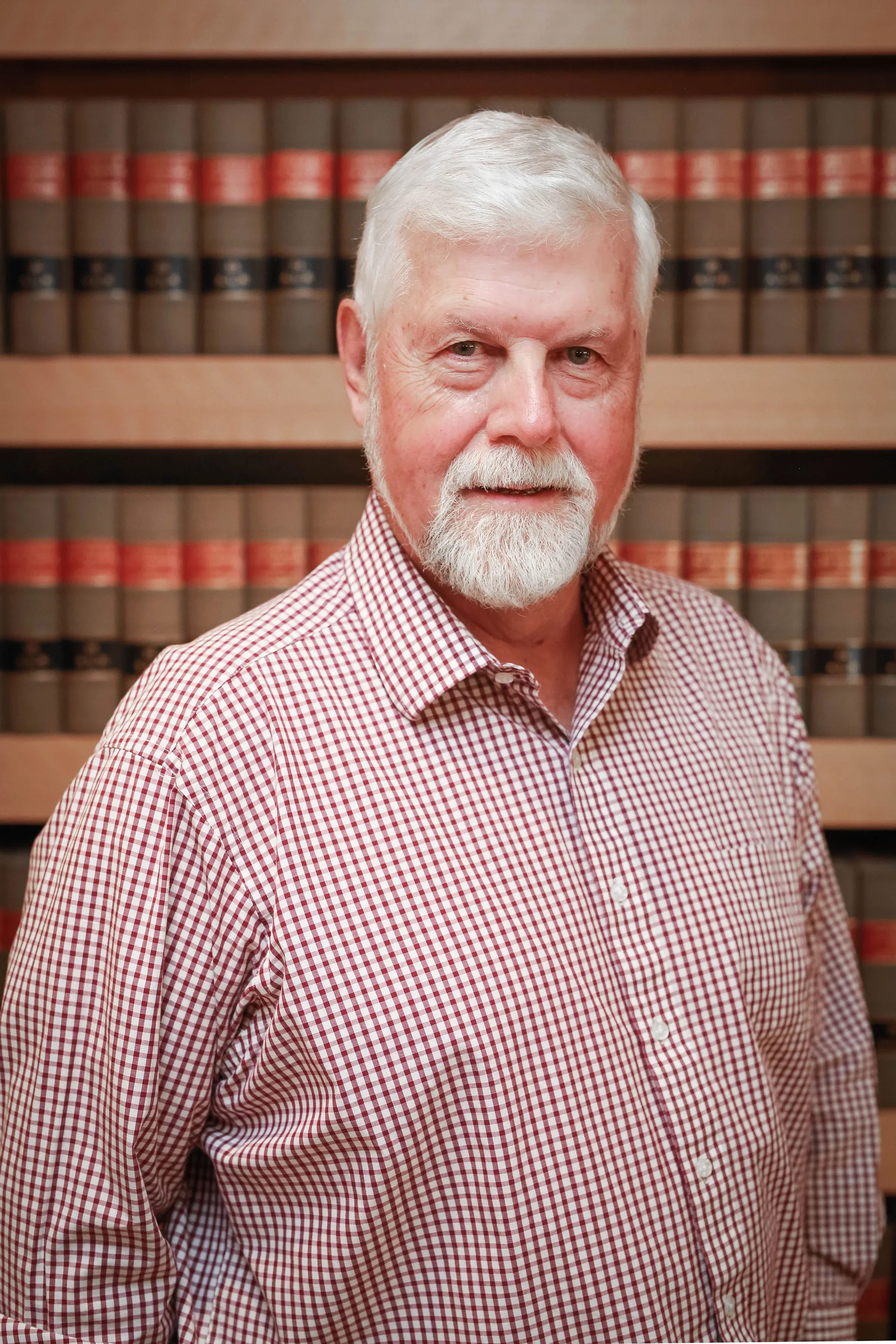 A professional headshot of a smiling man with white hair and a white beard, wearing a red and white plaid shirt, in front of bookshelves filled with legal texts.