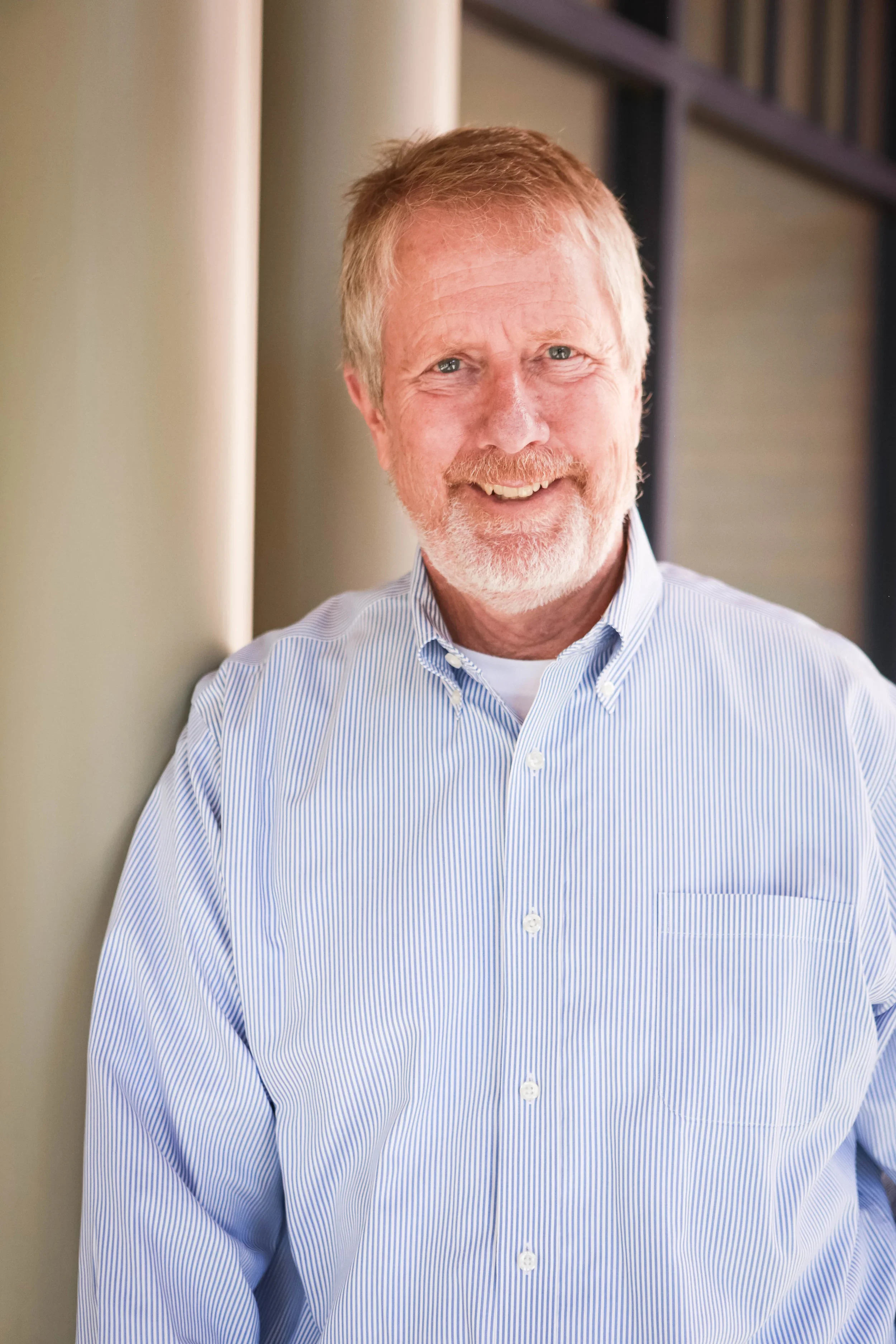 A professional headshot of a smiling man with a white beard and short blond hair, wearing a blue and white striped button-down shirt, leaning against a light-colored pillar.
