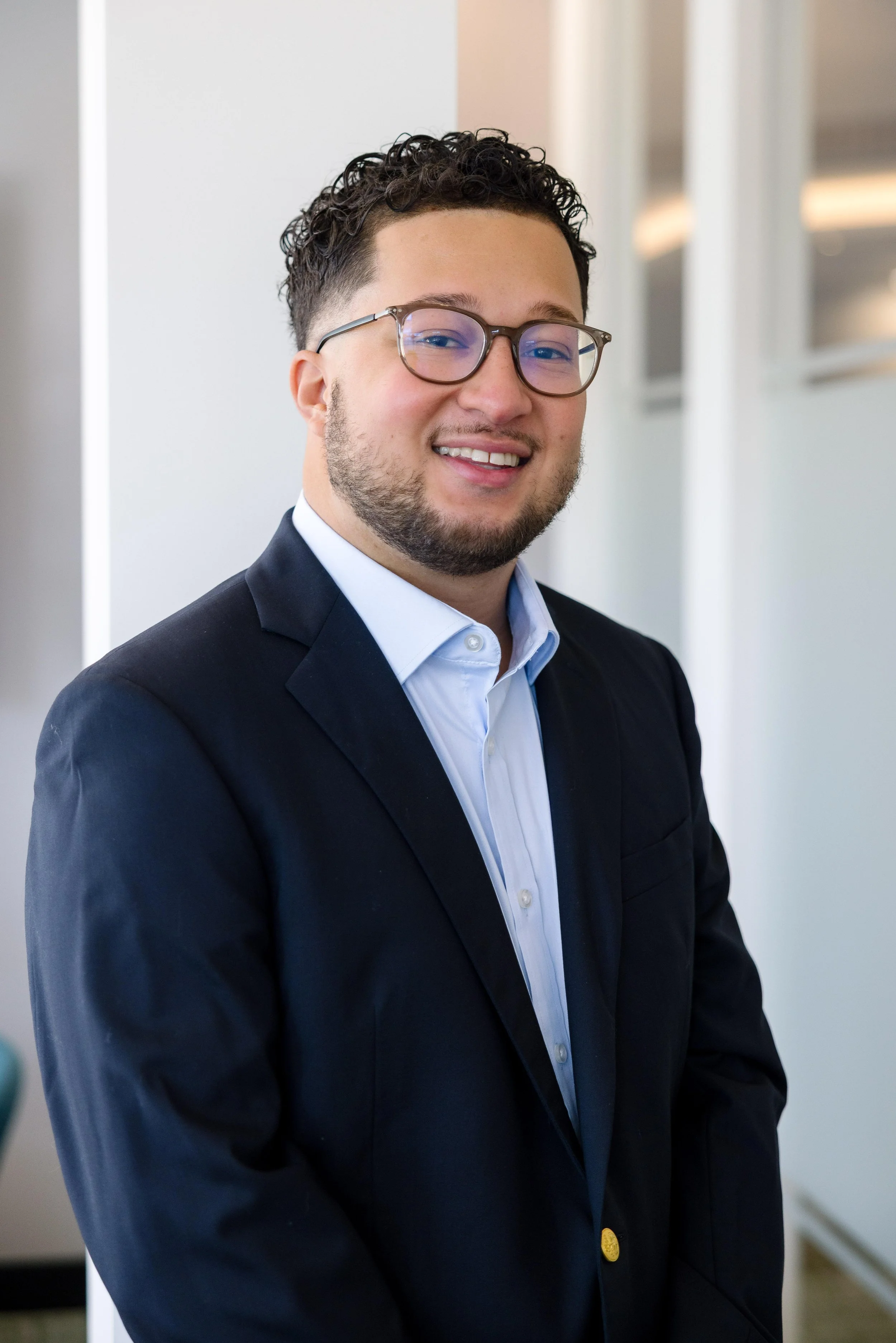 A professional headshot of a smiling man with glasses, dark hair, and a beard, wearing a navy blue blazer and a light blue dress shirt, standing in a brightly lit modern office.